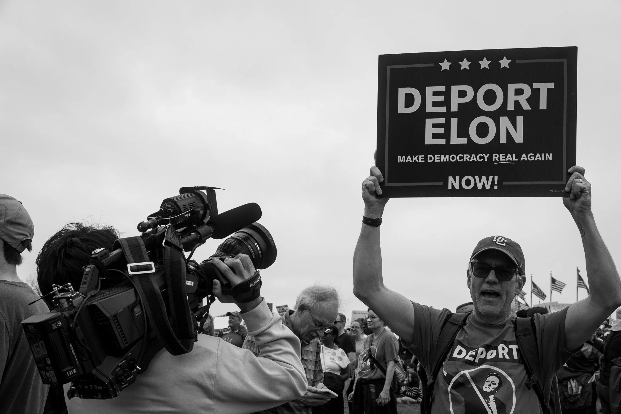 A man wearing sunglasses and a cap holding a sign that says 'Deport Elon' with additional text 'Make Democracy Real Again' and 'Now!', during a protest or rally. There are other people and flags in the background, and a cameraman filming the scene.