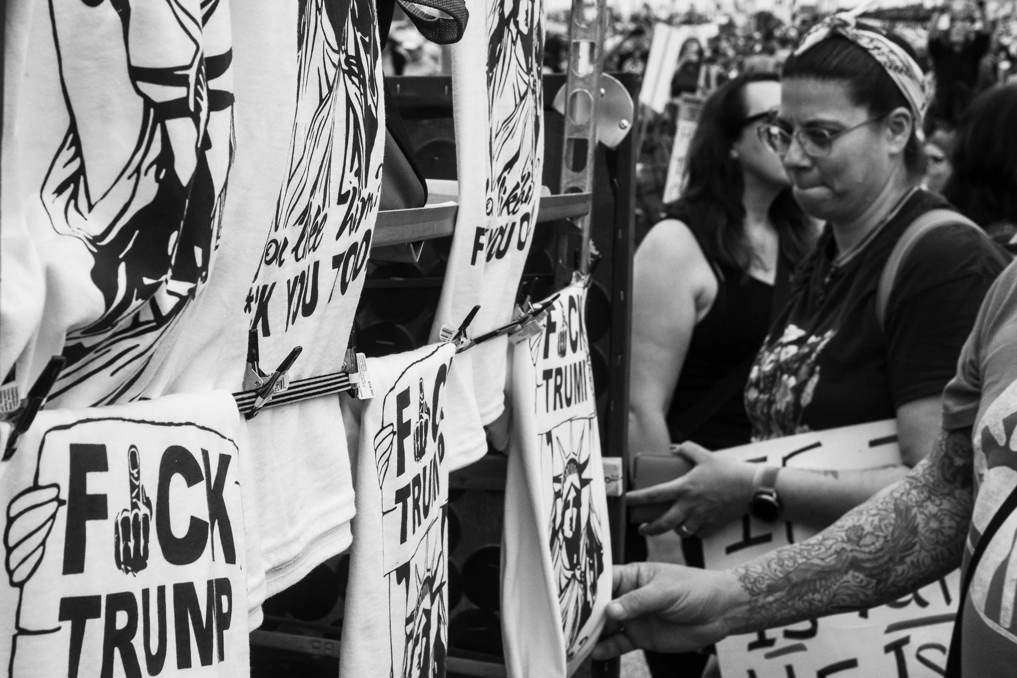 People browsing T-shirts with political messages at an outdoor booth, including one with a graphic of a hand making a middle finger and the phrase 'F**k Trump'.