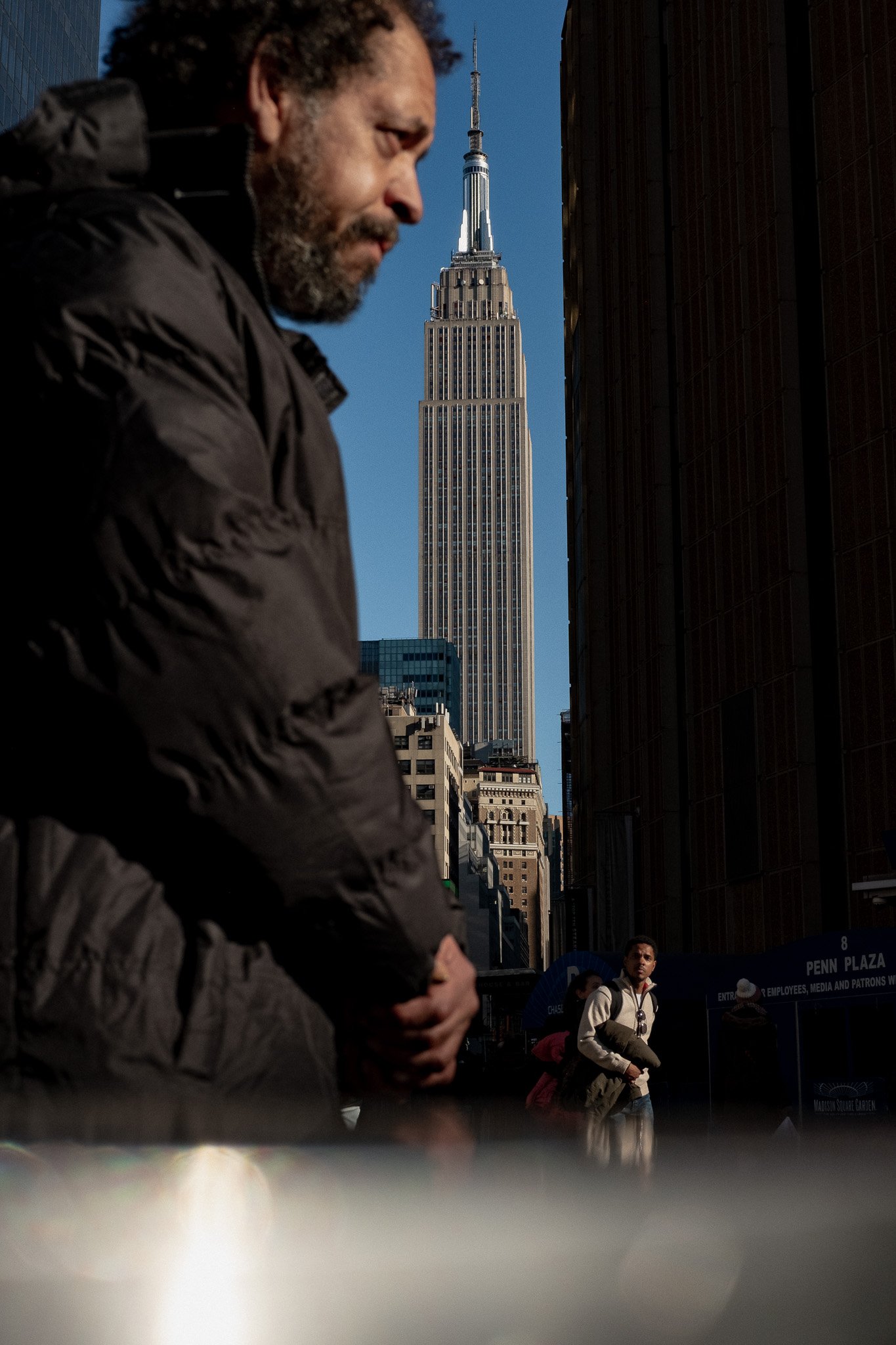 Street photo of A man with a beard in a dark jacket standing outdoors in front of the Empire State Building in New York City, with a few other people visible in the background.