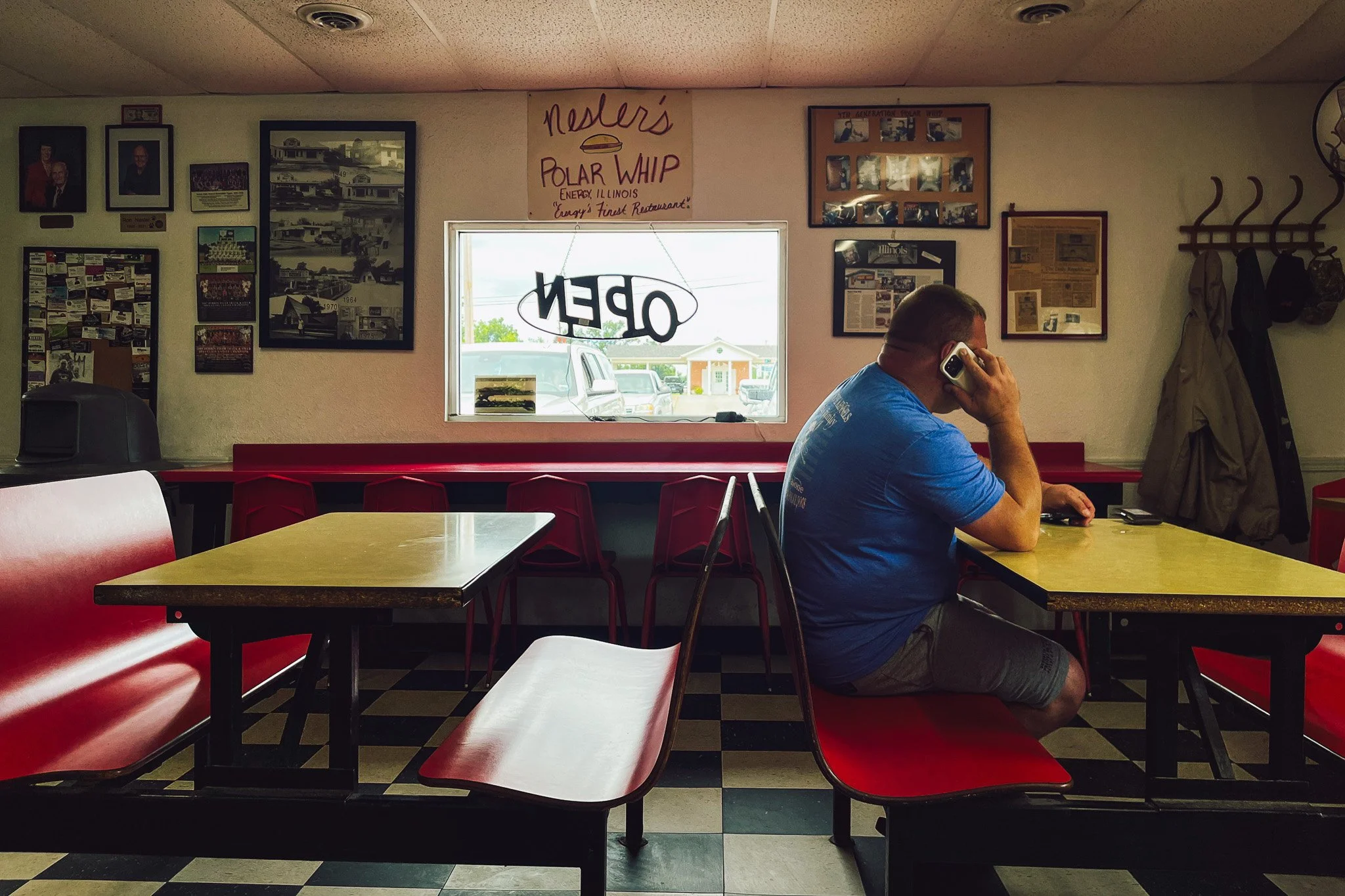 A man in a blue T-shirt and shorts sitting at a table inside a diner, talking on a cellphone. The interior has checkered black and white floors, red booths, and walls decorated with photos, posters, and a window with an 'OPEN' sign hanging. Outside, 