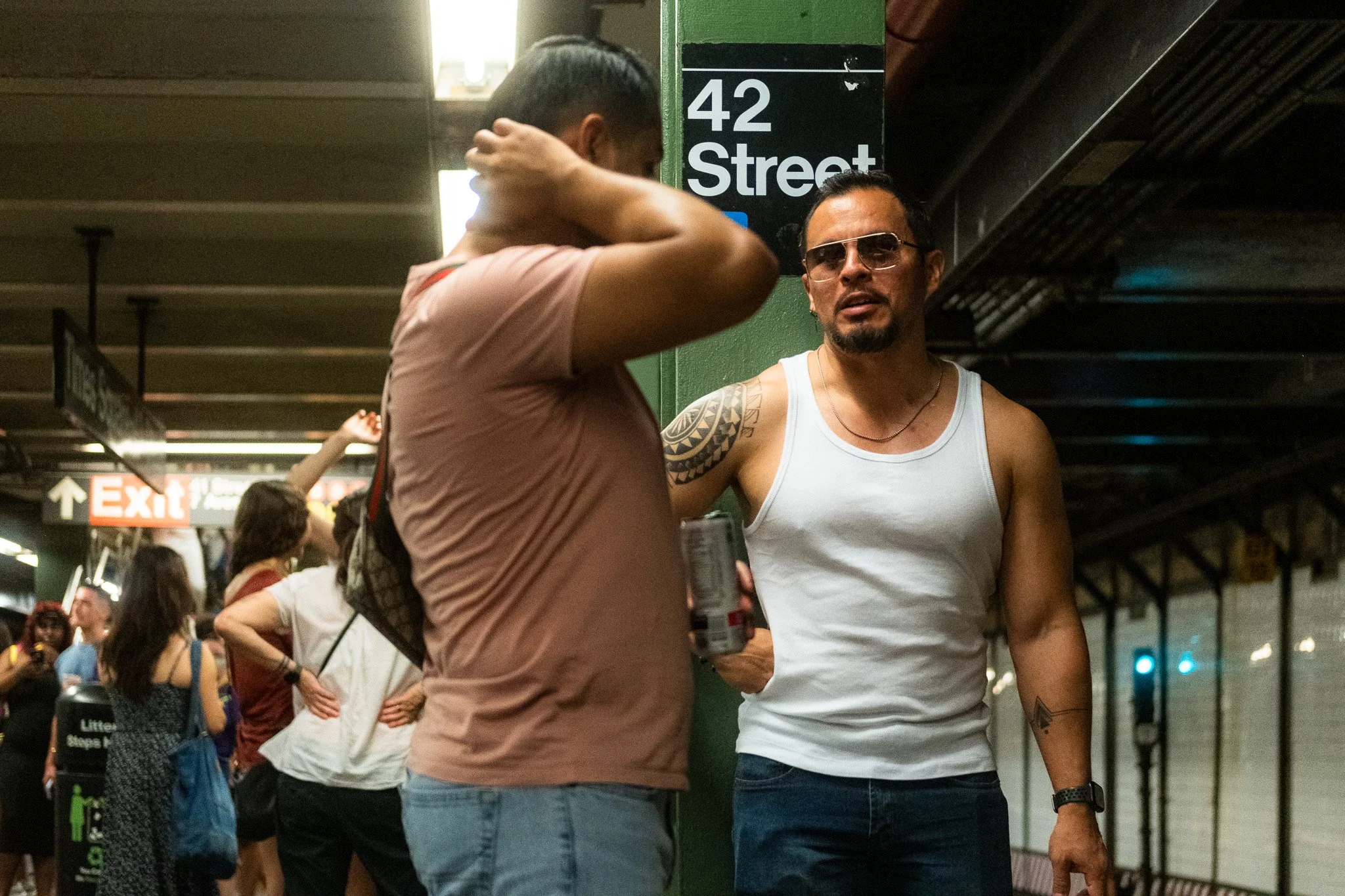 Street photograph of two men are talking in a subway station, one wearing sunglasses and a white tank top, the other with a pink shirt, near a green column with a street sign reading '42 Street'. There are several people in the background, some with 
