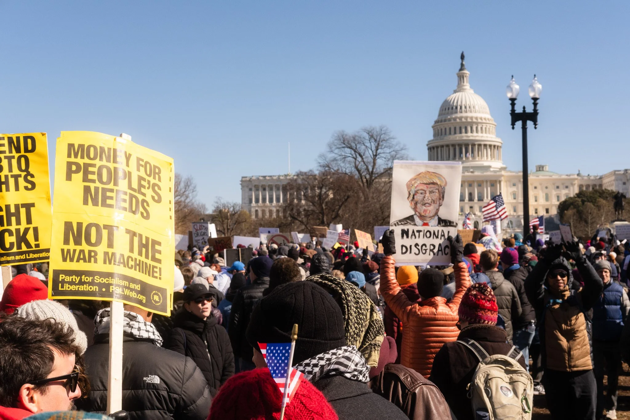Crowd of protesters gathered in front of the United States Capitol building, holding signs and flags, with clear blue skies above.