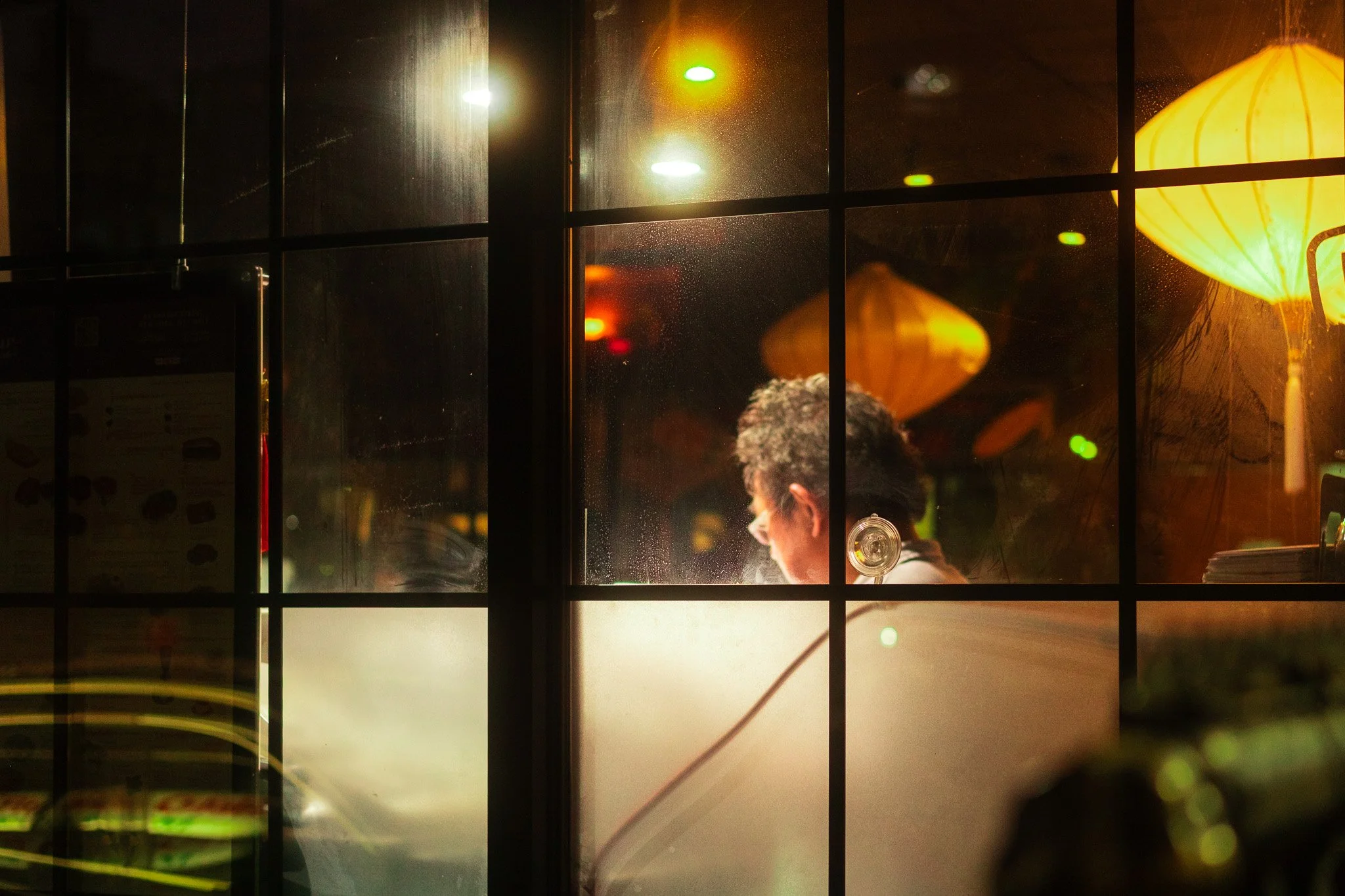 Street photograph of a man working inside a dimly lit restaurant, seen through a window with a grid pattern, with hanging lanterns providing warm light.