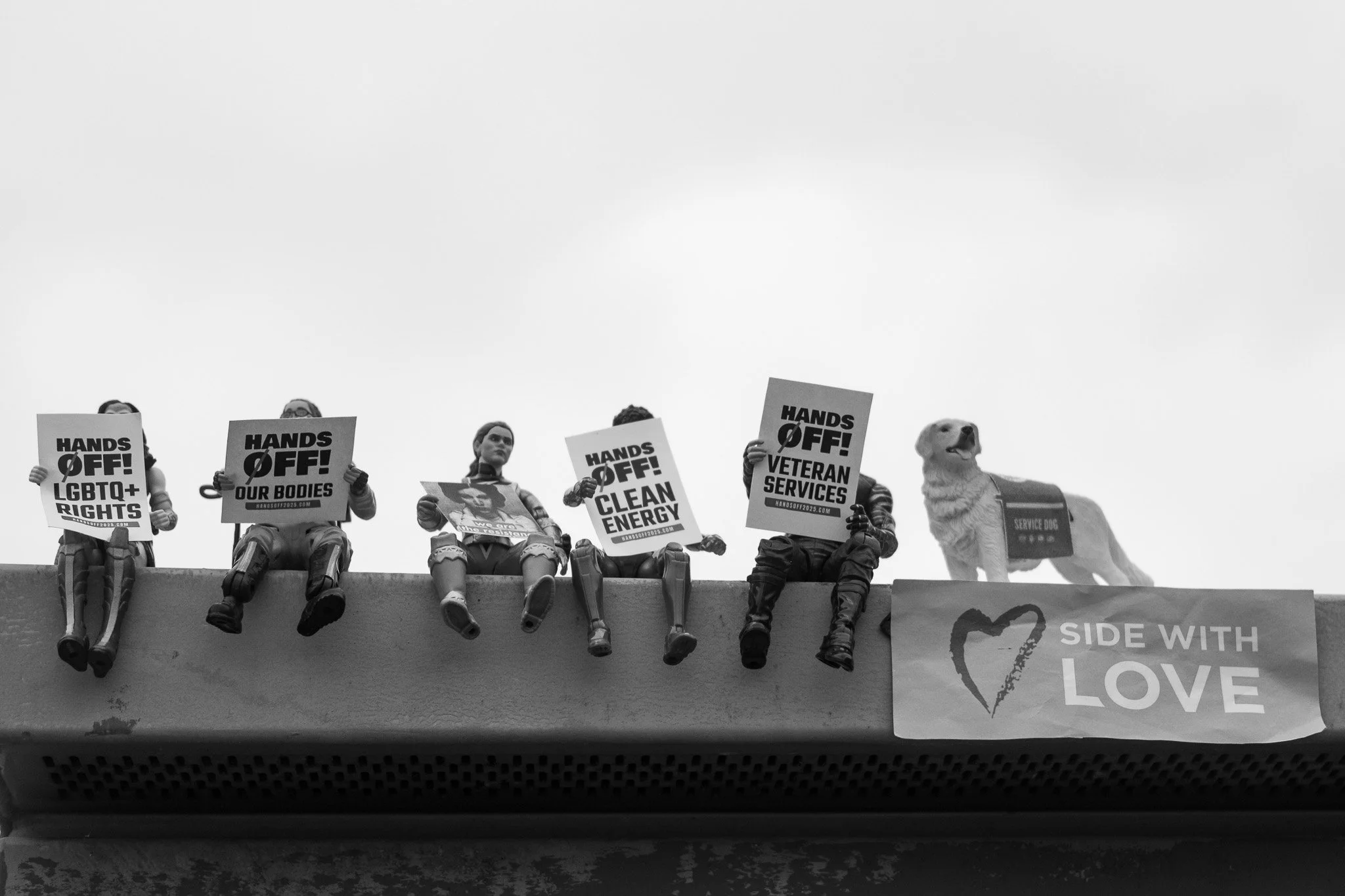 Five toy figures sitting on a ledge holding protest signs with messages like "HANDS OFF! LGBTQ+ RIGHTS," "HANDS OFF! OUR BODIES," "HANDS OFF! CLEAN ENERGY," and "HANDS OFF! VETERAN SERVICES." There is a dog with a service dog vest on the far right an