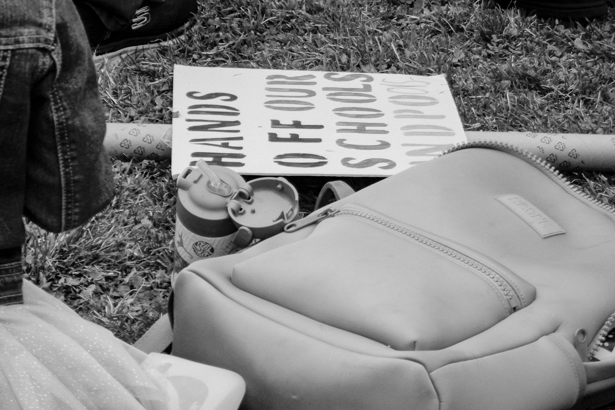 A protest sign on the ground that reads 'THANKS GOD FOR OUR SCHOOLS' with a Starbucks drink and a backpack nearby, on a patch of grass.