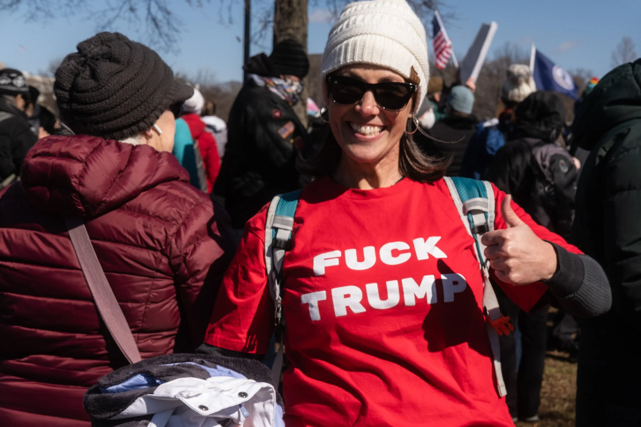 A woman at a protest or rally wearing a red shirt that says 'F*** TRUMP,' smiling, with sunglasses and a white knit hat, giving a thumbs-up, with a crowd in the background holding flags.