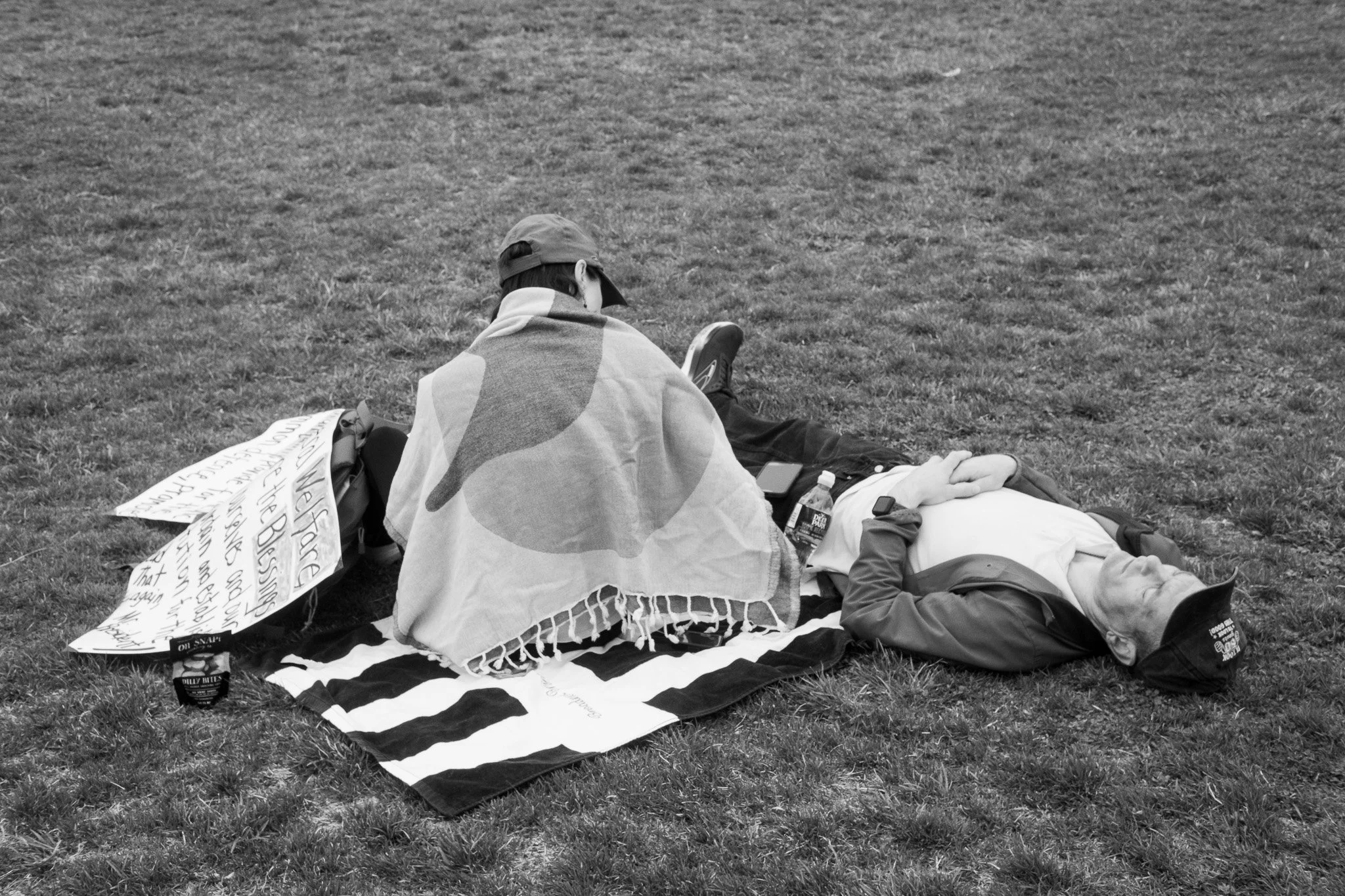 A person sitting on a striped blanket on the grass with another person lying down, holding hands. The sitting person has a towel draped over their shoulders. Items around include signs, water bottles, a phone, and snacks.