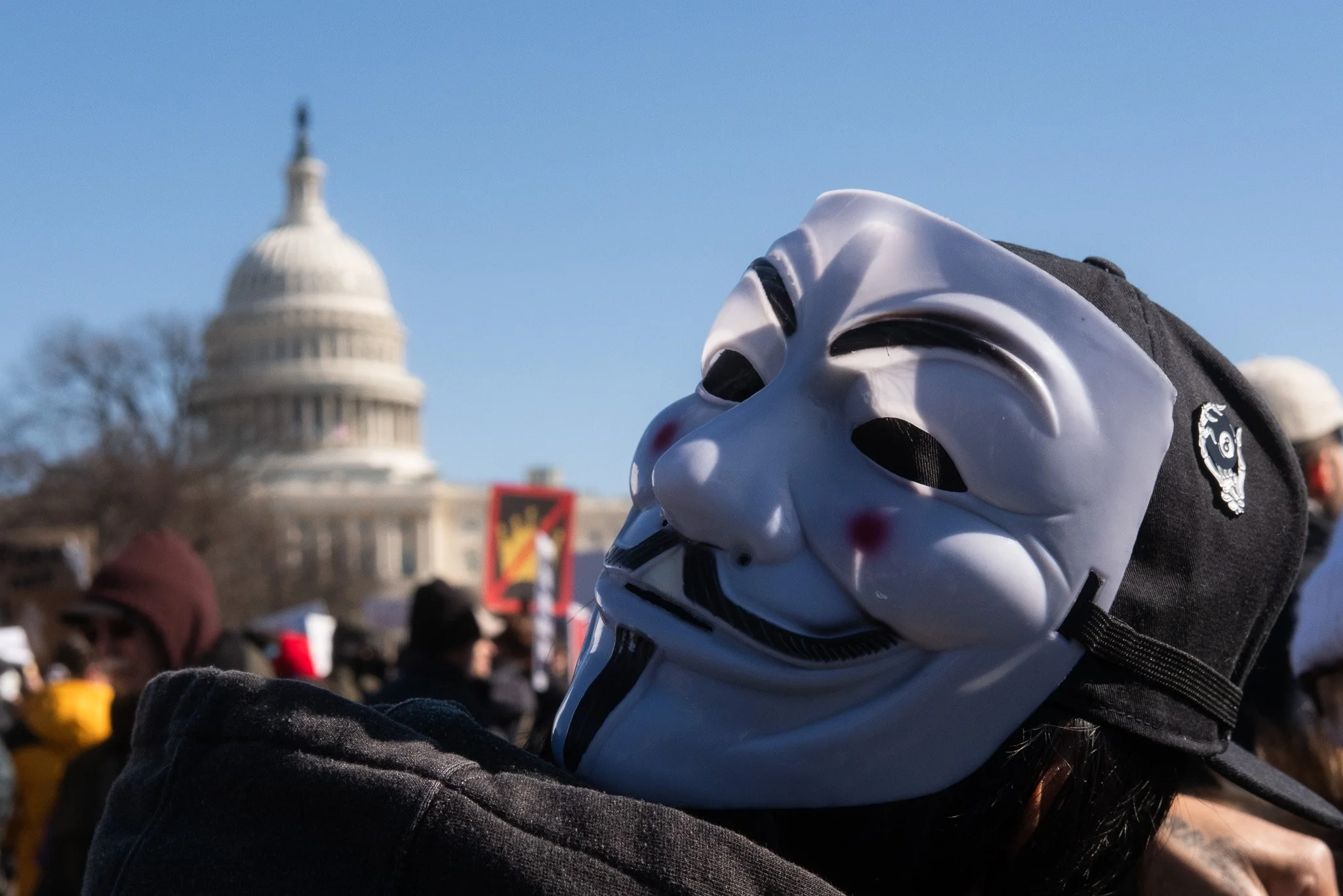 Person wearing a Guy Fawkes mask in front of the U.S. Capitol building during a protest or rally.