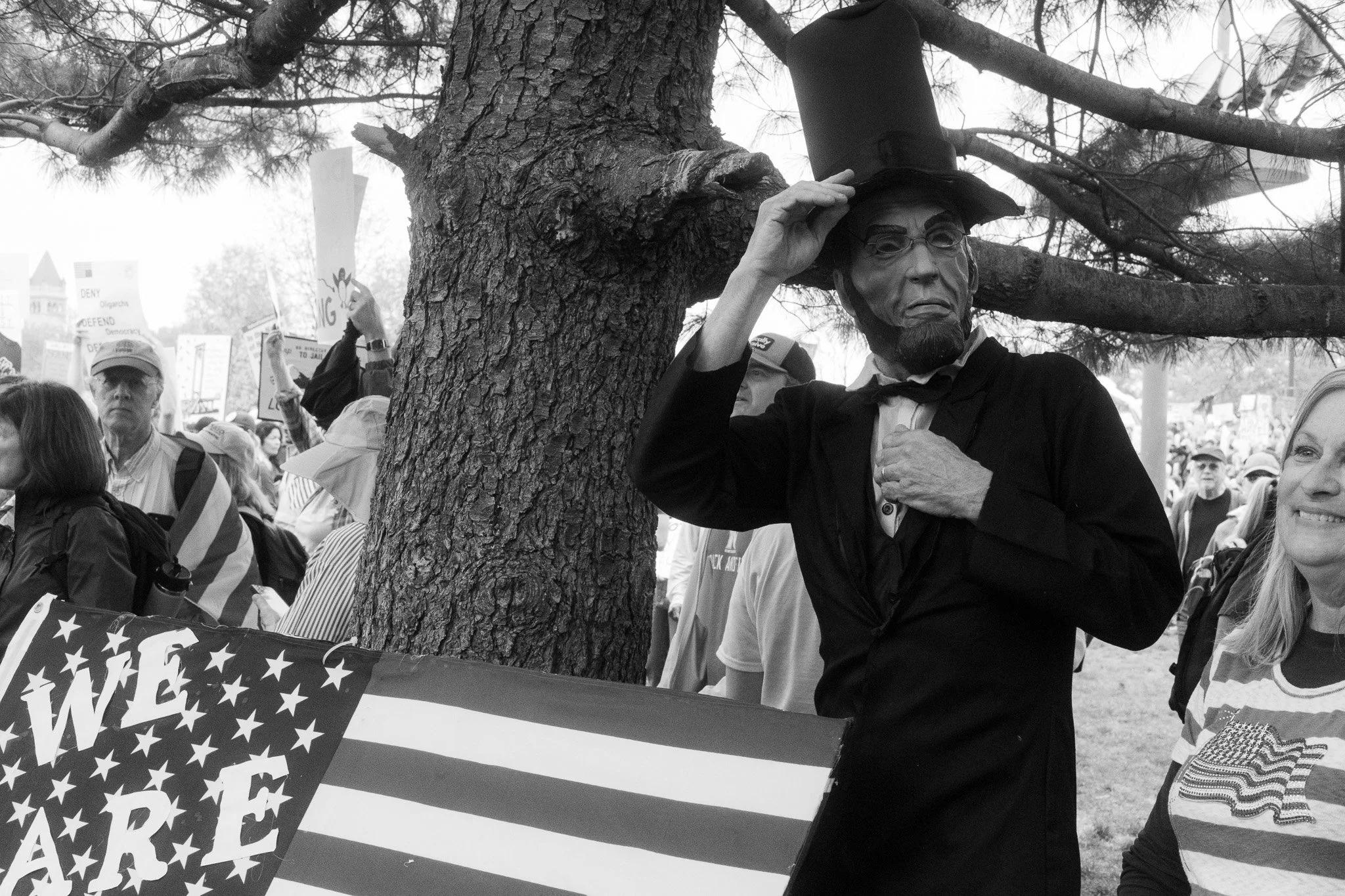 A person dressed as Abraham Lincoln, wearing a tall black hat, glasses, and a beard, is standing outdoors among a crowd, leaning against a tree, with one hand on their chest and the other adjusting their hat. The crowd holds signs and flags, and a si