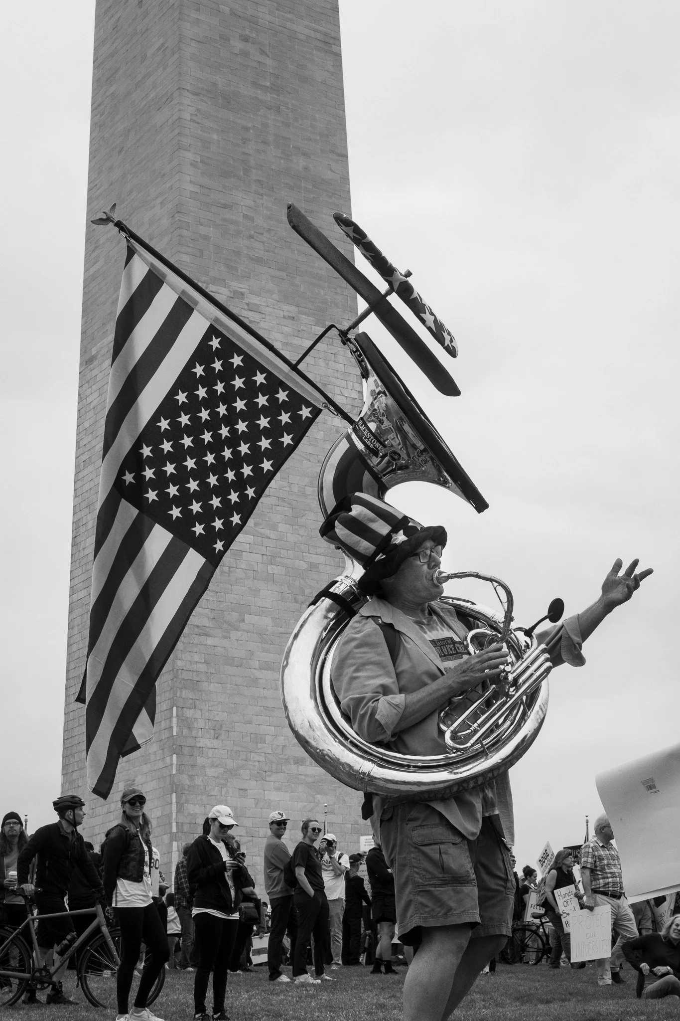 A person playing a saxophone while walking in a protest or rally, with a large American flag, a microphone, and a bicycle flag attached to their equipment. The person is wearing glasses and a hat, and there are multiple people in the background.