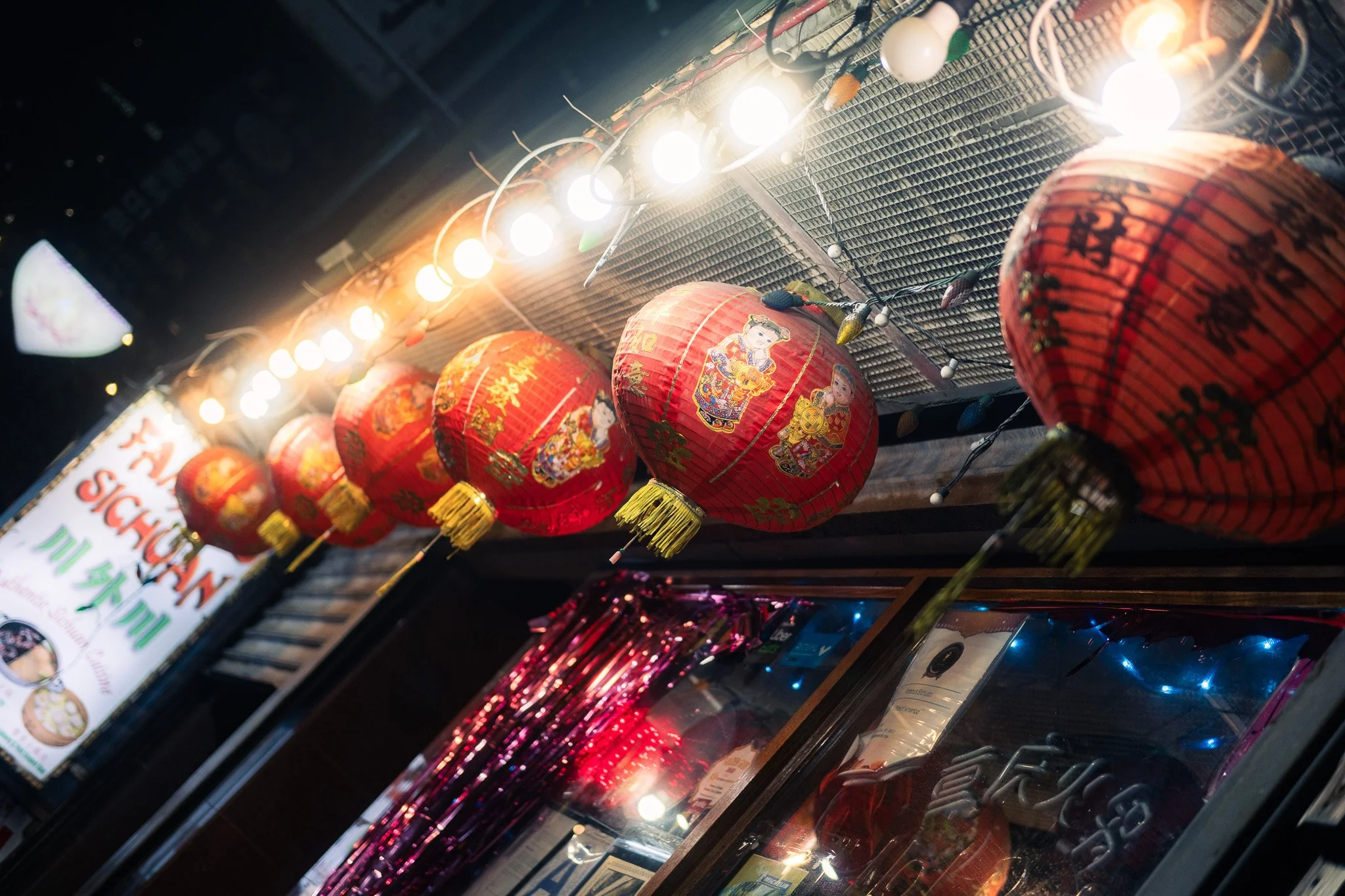 Red Chinese lanterns hanging outdoors with bright lights above, decorated with Chinese characters and images of children in traditional attire, reflected in a nearby glass window.