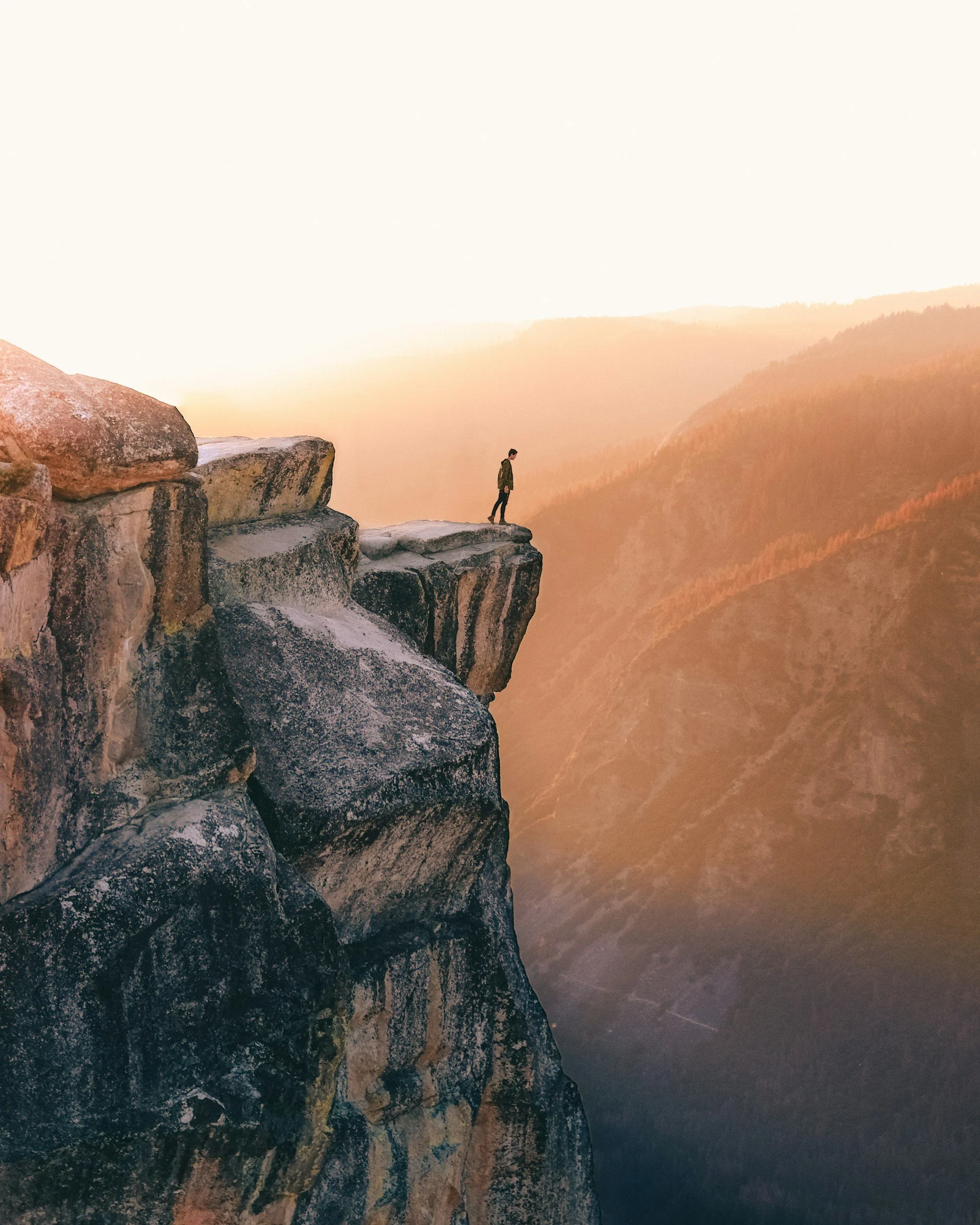 Person standing near the edge of a rocky cliff overlooking a valley at sunset