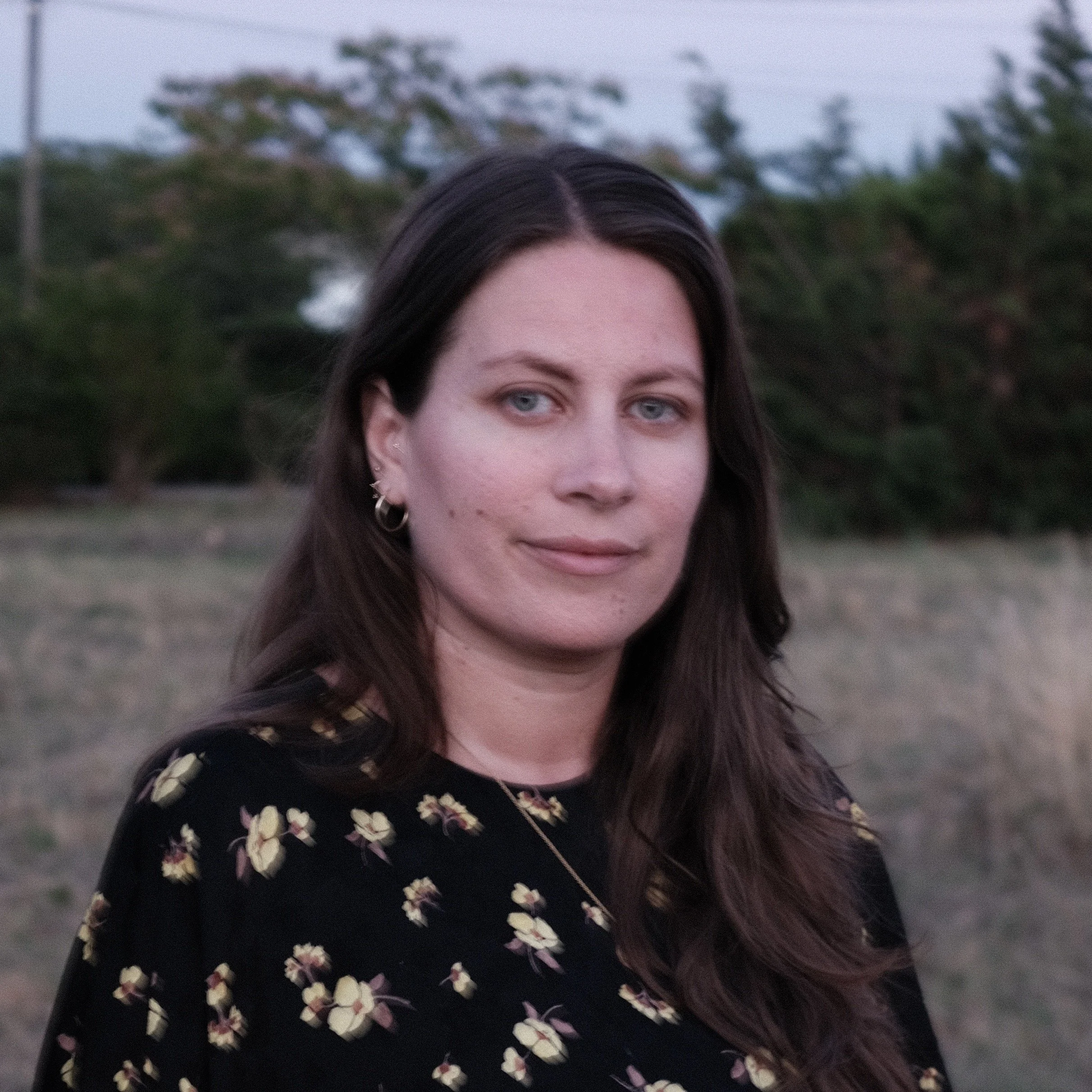 Close-up portrait of a female brunette in a field at dusk, with long brown hair, grey-green eyes, and a black and yellow floral-print dress.