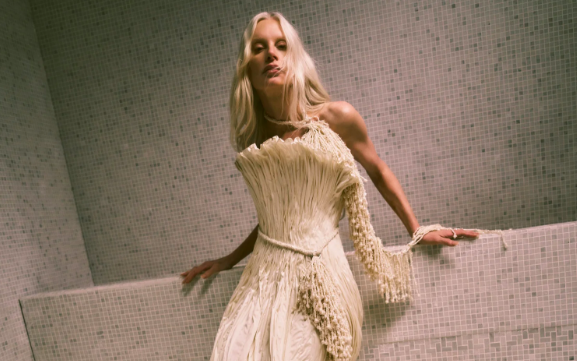 Model Kristen McMenamy with platinum-blonde hair, wearing a white strapless couture bridal gown by Danielle Frankel, propped up against a grey mosaic wall. The wedding dress is artfully textured, fans out at the neckline and has a cinched waist.