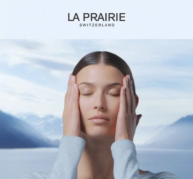 Serene image of a brunette woman applying skincare on her face with her eyes closed. Hazy Swiss mountains and a lake are in the background behind her, as well as cloudy blue skies.