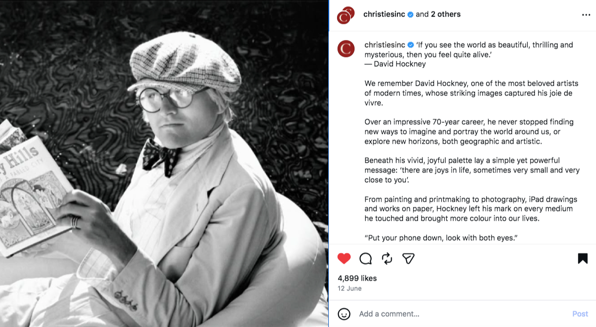 Vintage black and white photo of David Hockney wearing round glasses and a baker-boy cap and a suit while reclining in a chair, reading a newspaper.