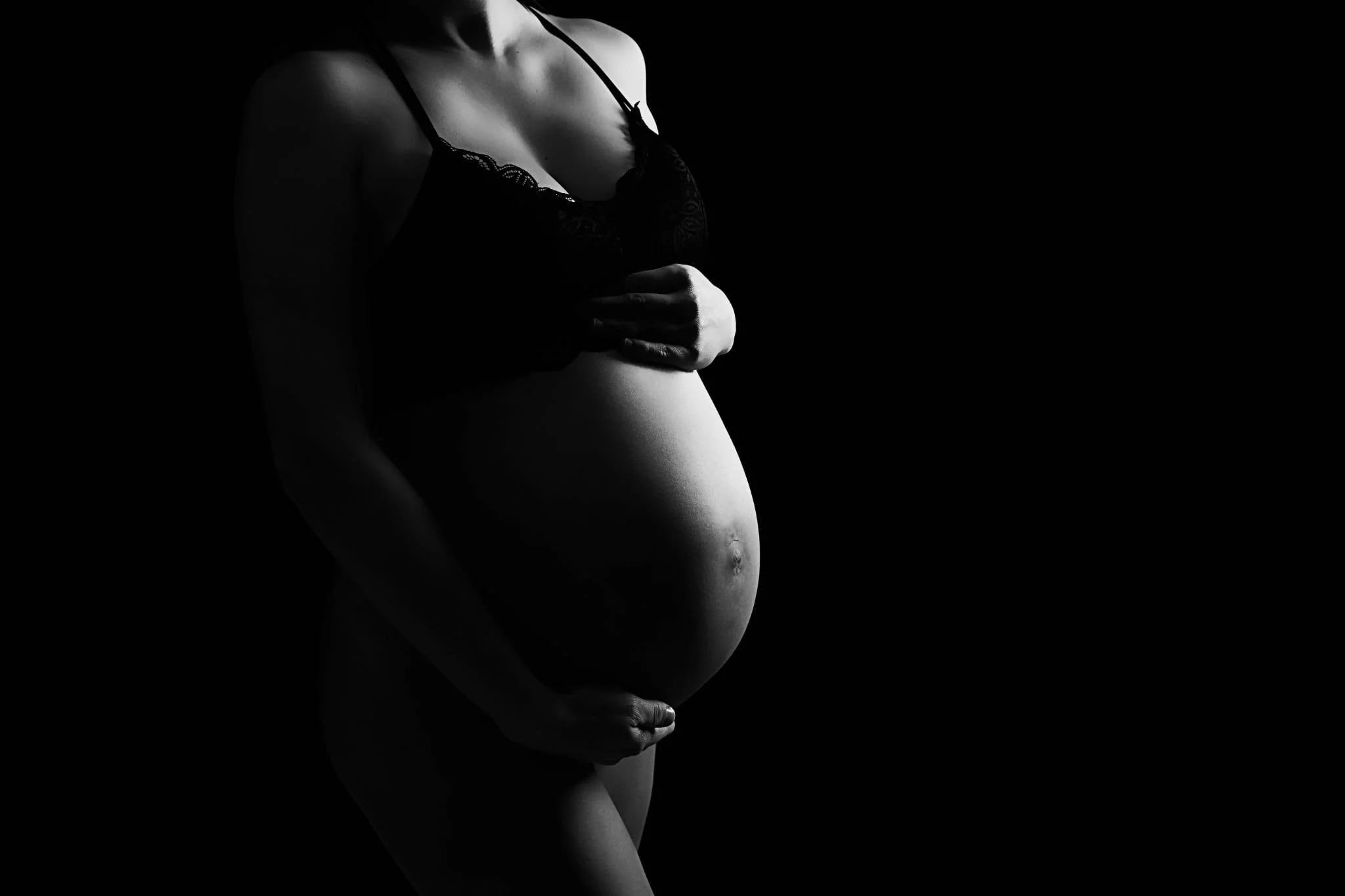 Black and white photo of a pregnant woman with her hand on her belly, wearing a lace bra, against a dark background.