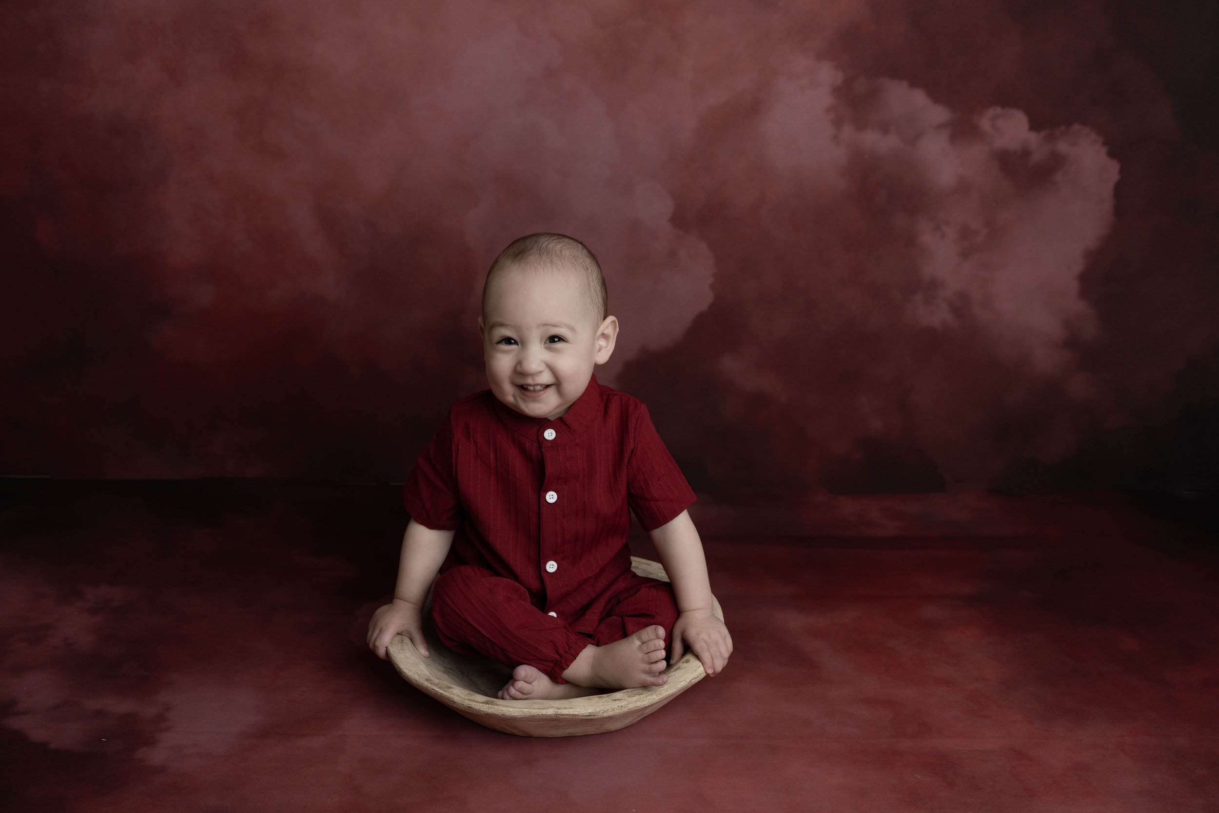 A smiling baby boy wearing a red outfit, sitting on a wooden bowl with his legs crossed, against a dark burgundy and purple cloudy background.