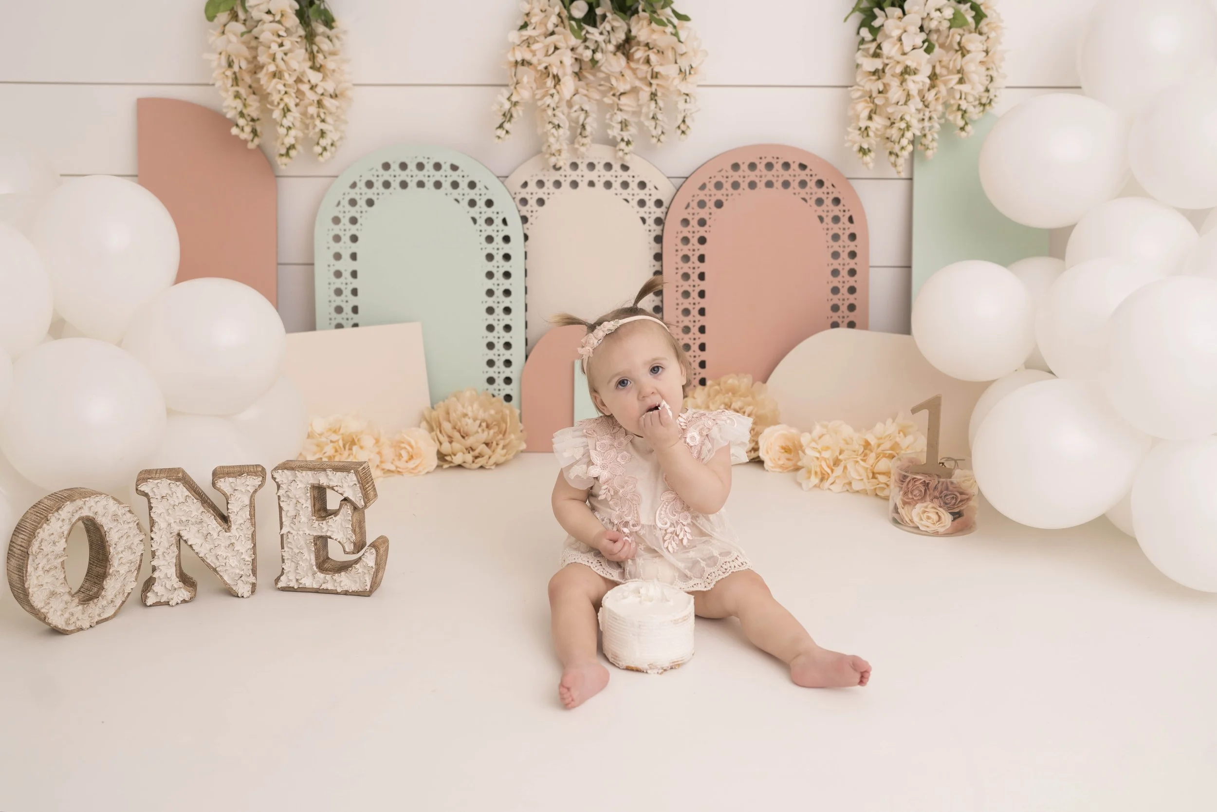 A young girl in a pink lace dress sitting among pastel colors, floral decorations, white balloons, and decorative letters spelling 'ONE' on a white background.