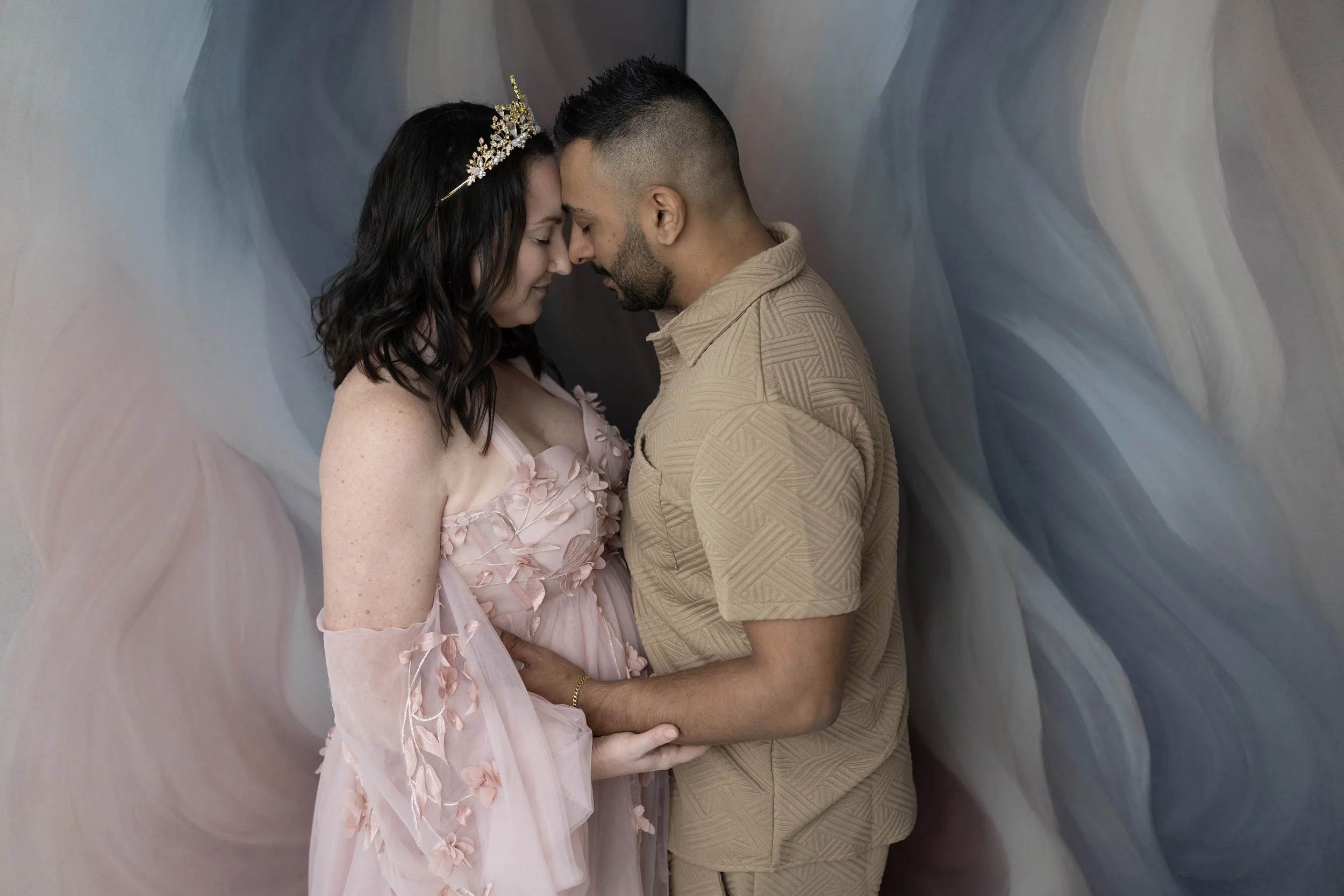 A couple standing close together with foreheads touching, eyes closed, and holding hands. The woman is wearing a pink dress with floral details and a tiara, while the man is in a beige patterned shirt. They appear to be in an intimate, romantic moment against a soft, abstract background.