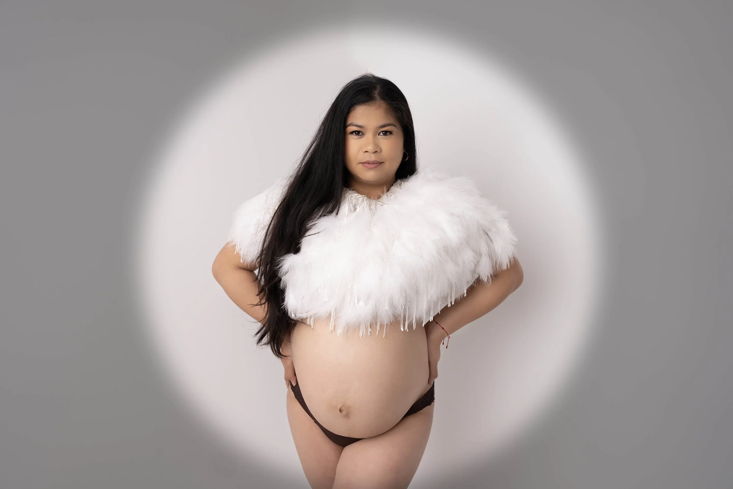 Woman with long dark hair wearing a white fluffy shawl and black lingerie posing against a gray background.