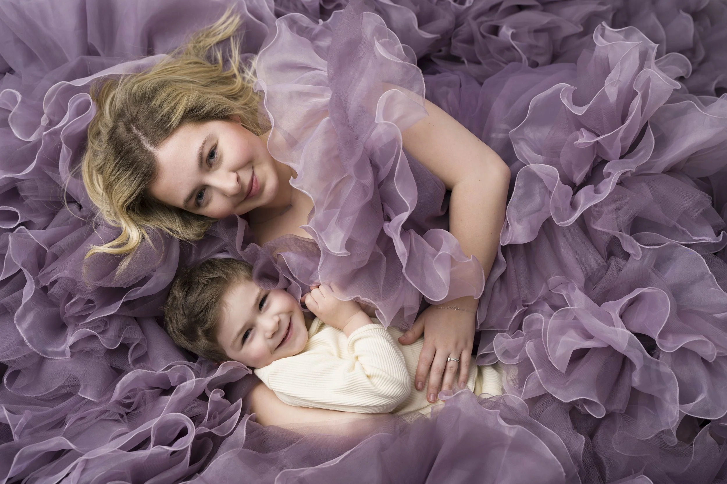 A woman with blonde hair lying on a bed surrounded by purple ruffled fabric, smiling and holding two young children, one boy and one girl, all smiling.