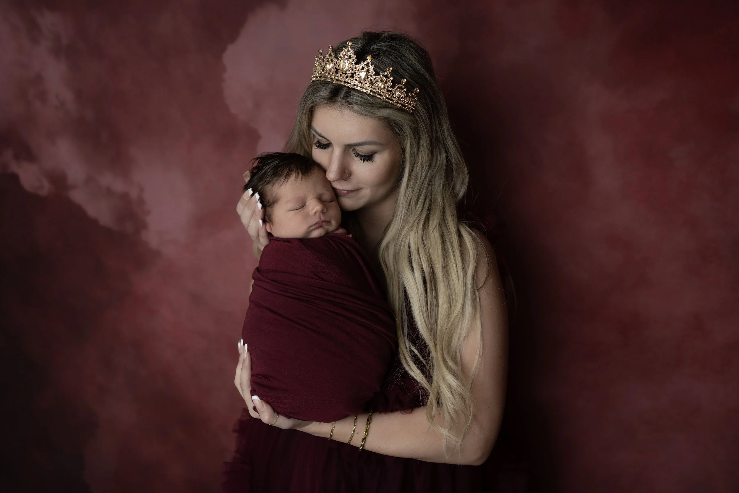 A woman with long blonde hair wearing a tiara holds a sleeping baby wrapped in a burgundy blanket against a dark red and purple background.