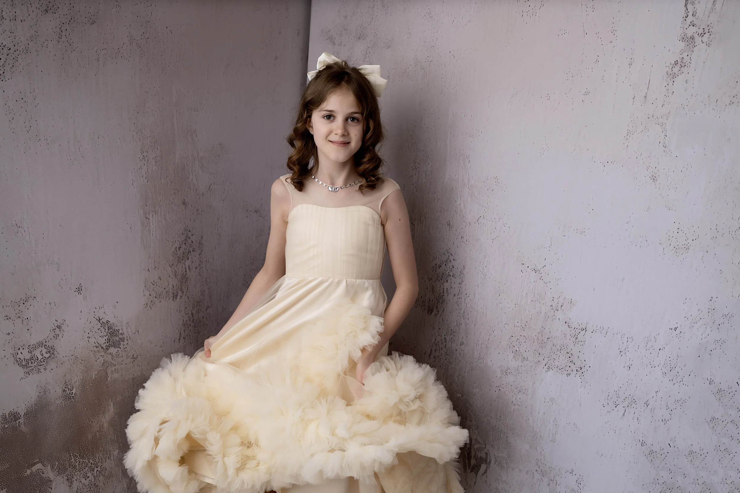 A young girl in a cream-colored dress with a fitted bodice and a fluffy, ruffled skirt, standing against a textured gray wall. She has wavy brown hair, a white bow headband, a necklace, and is smiling softly.