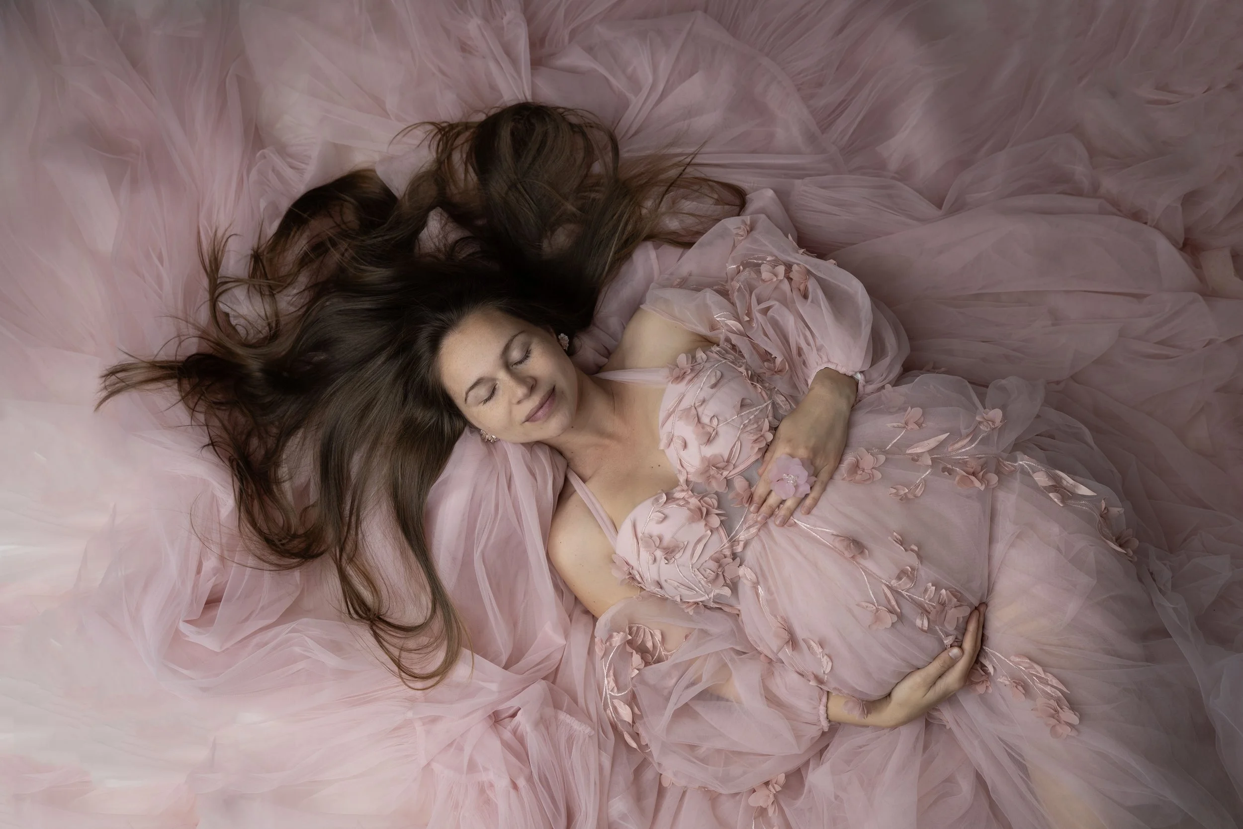 Two women lying on pink, ruffled fabric, both wearing matching pink dresses with floral embellishments and with their eyes closed.