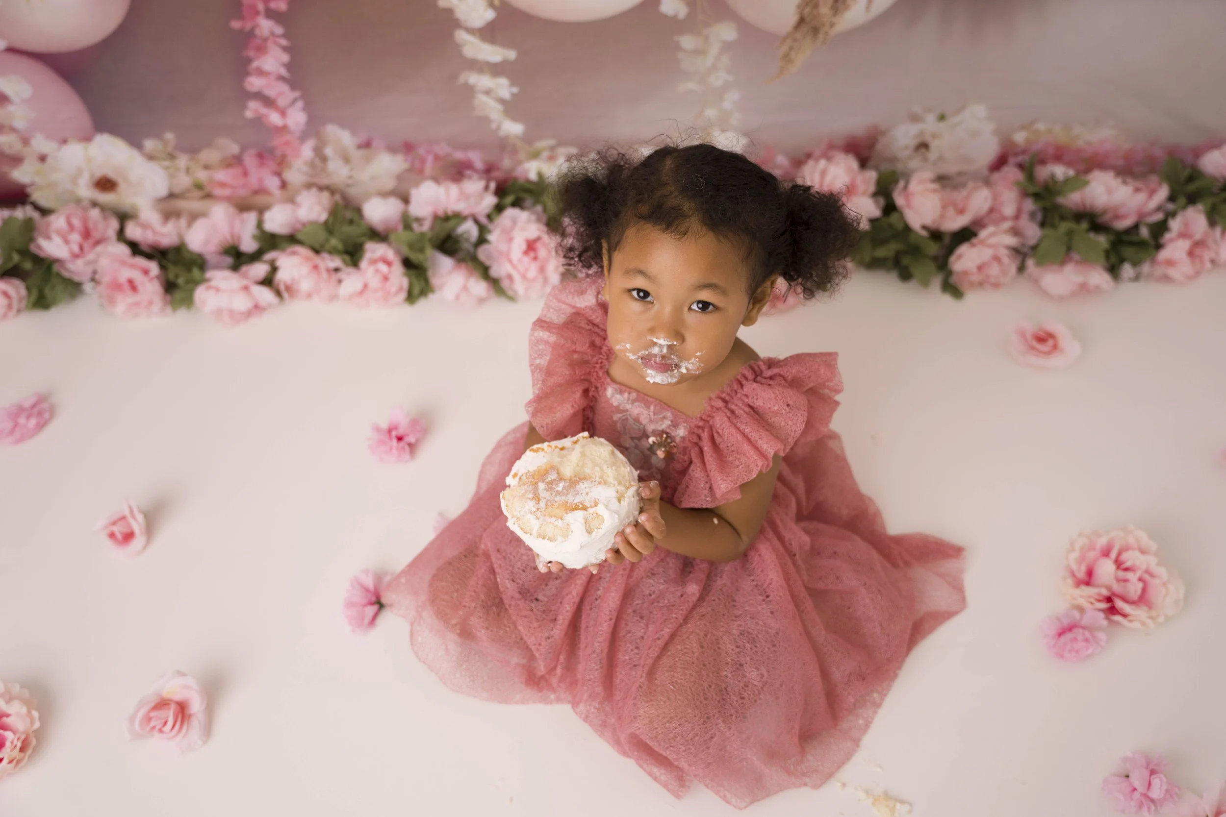 A young girl with curly hair and a pink dress sitting on a pink surface, holding a cupcake with white frosting, surrounded by pink and white flowers.