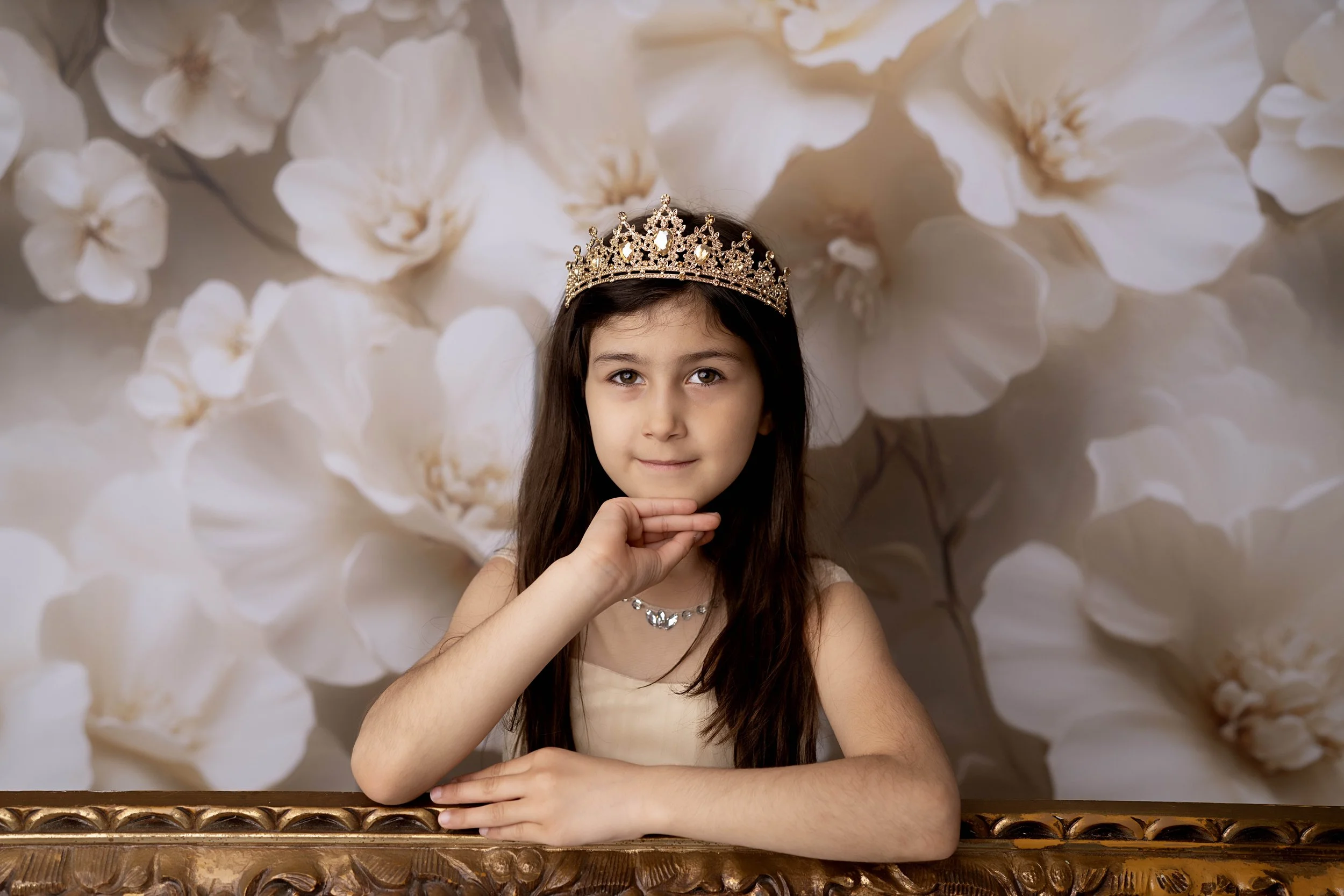 A young girl with long brown hair wearing a tiara and a necklace, resting her chin on her hand, sitting behind a wooden table, with a backdrop of white flowers.