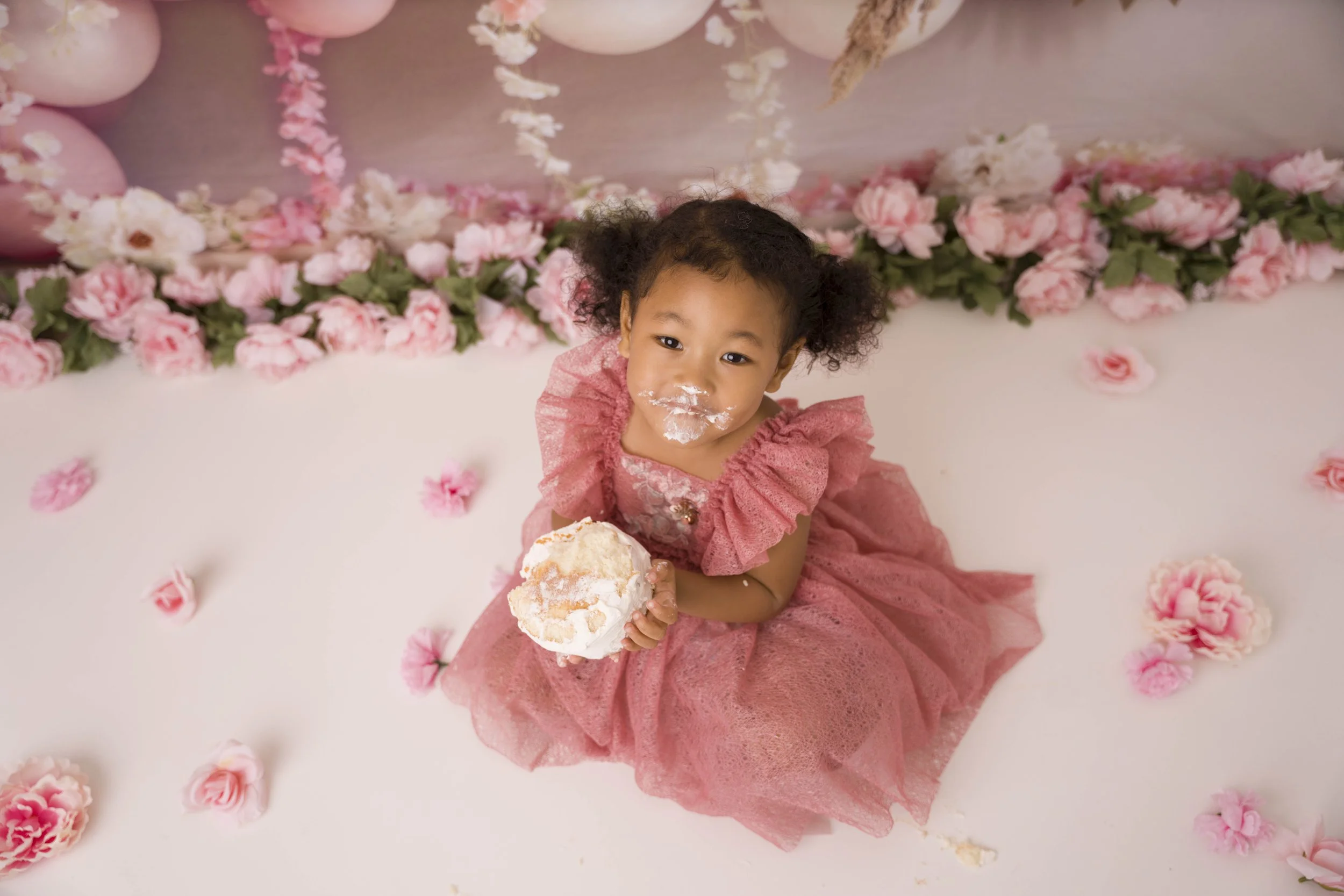 A young girl with curly hair wearing a pink dress sitting on a white surface decorated with pink roses and balloons. She is holding a large piece of cake, with some cake and frosting on her face.
