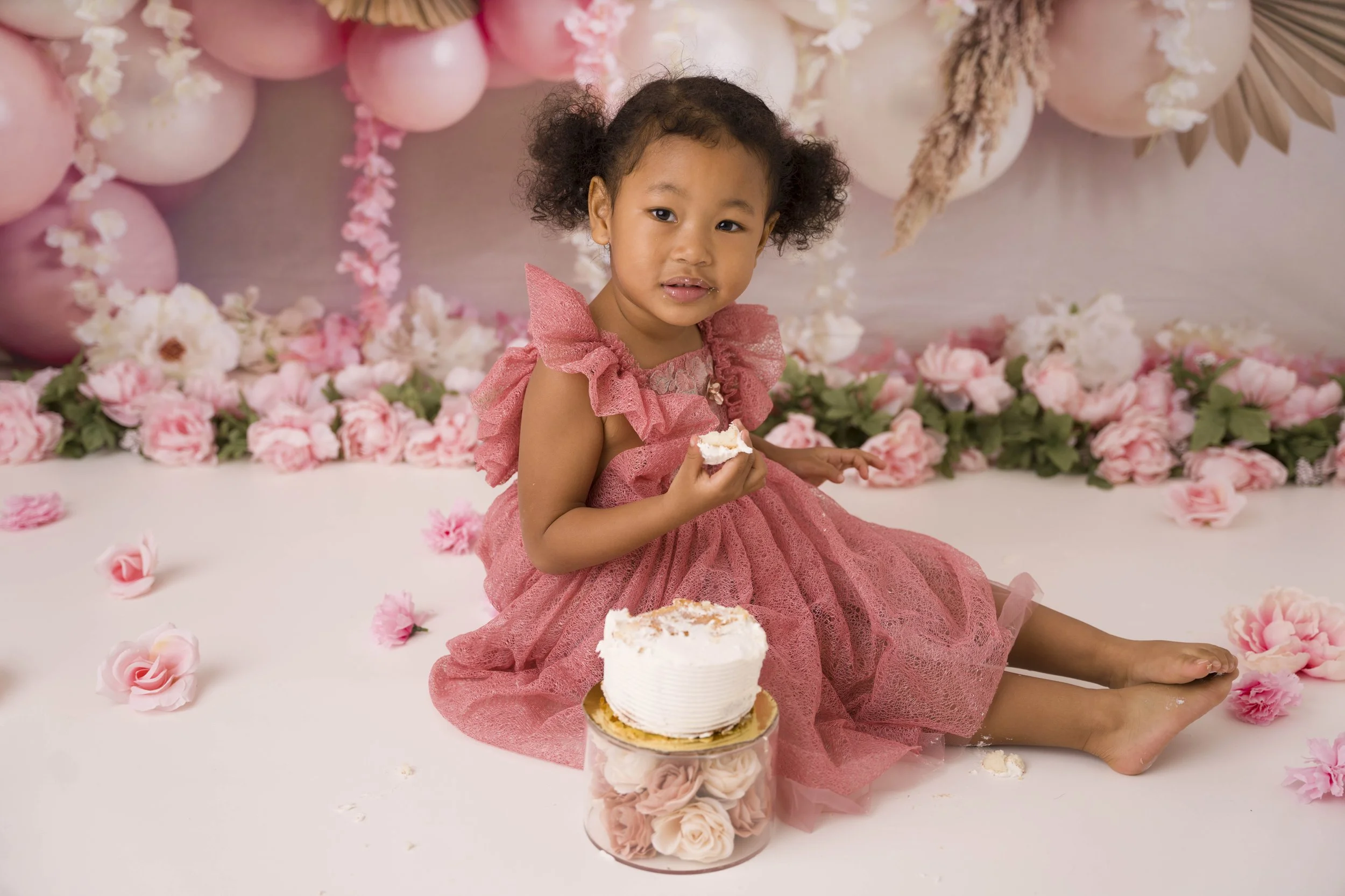A young girl wearing a pink dress with ruffles sitting in front of pink paper balloons and flowers, holding a piece of cake, with a decorated pink cake in front of her and flowers scattered on the table.