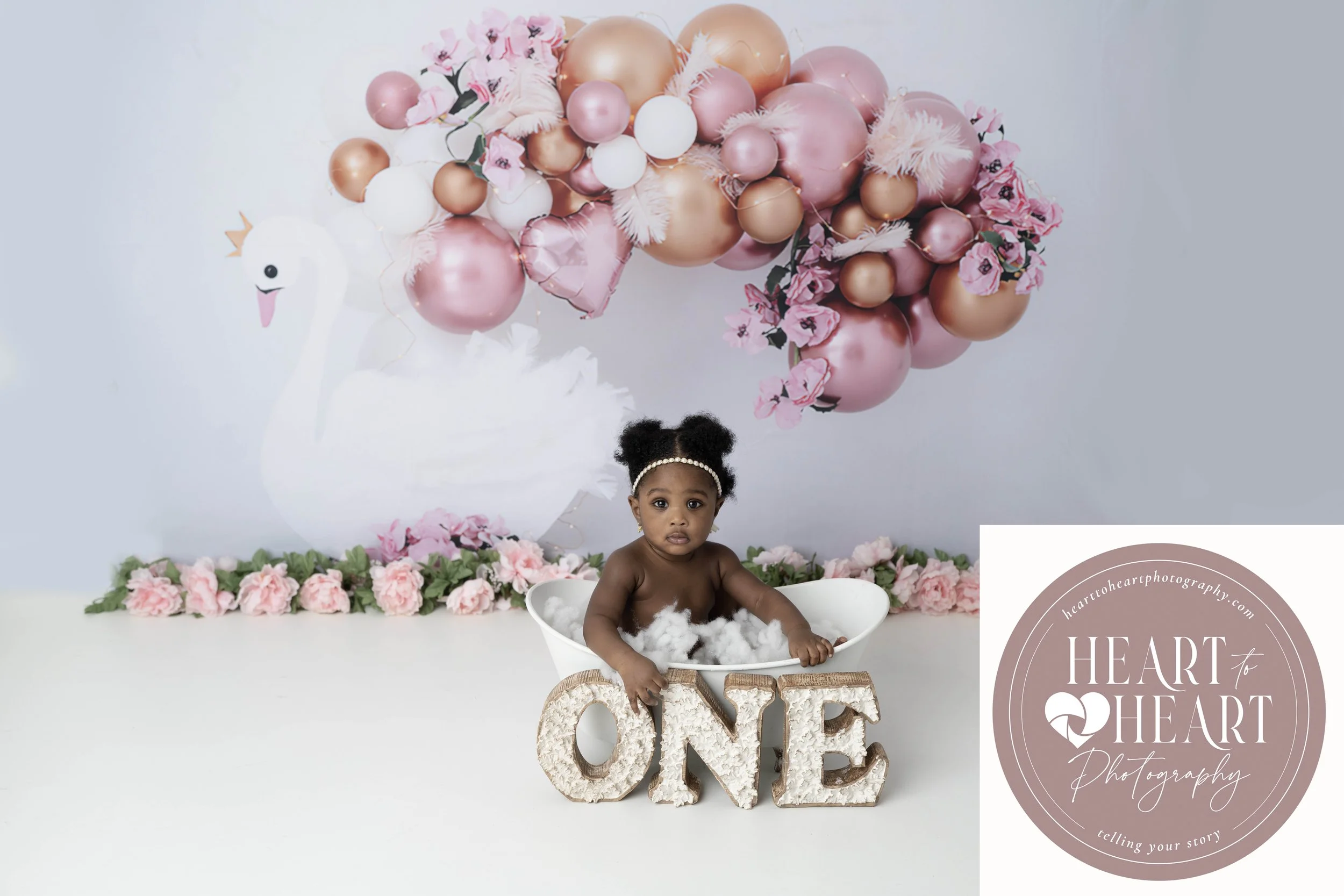 A young girl sitting in a bathtub filled with cotton, with a backdrop of pink and gold balloons and flowers, and a decorative "LOVE" sign in front of her.