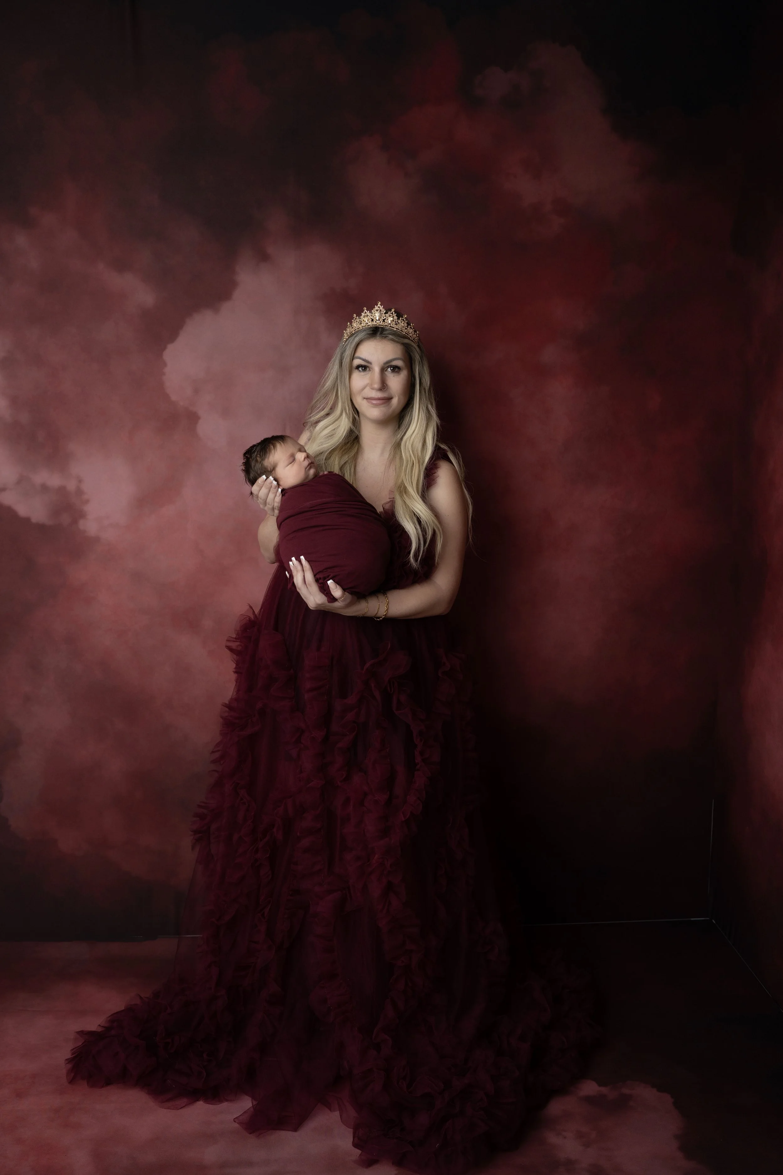 A woman wearing a crown and a burgundy gown holding a newborn baby wrapped in a matching burgundy blanket, standing against a red and black marbled background.