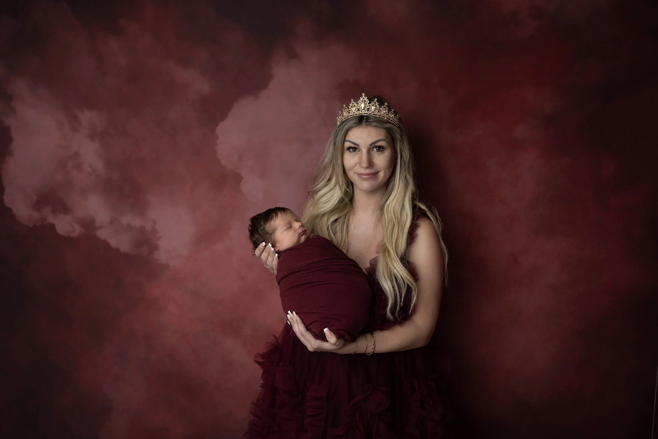 A woman with long blonde hair wearing a gold crown, holding a sleeping baby wrapped in a burgundy blanket, against a dark red and pink cloudy background.