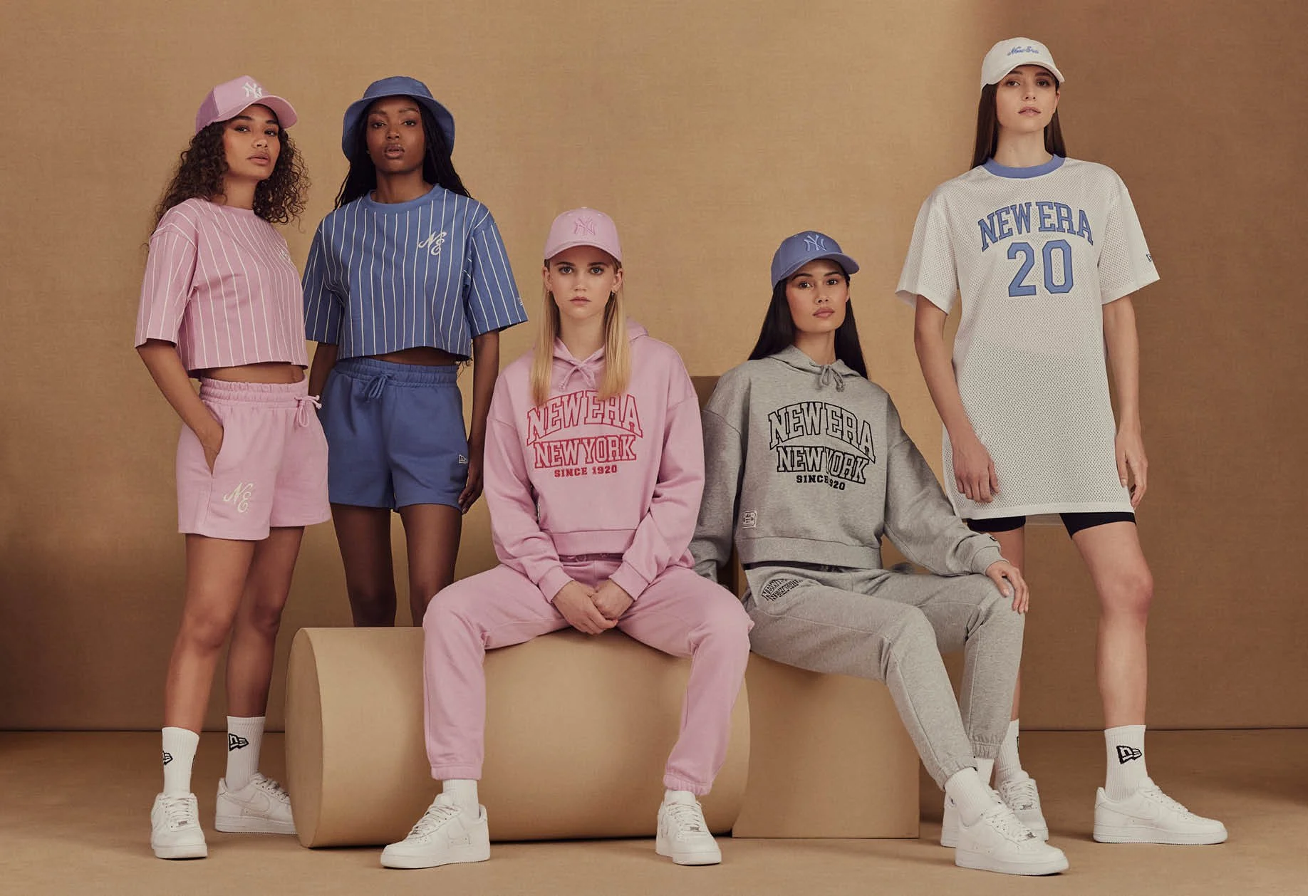 Group of six young women modeling casual sportswear and baseball caps in a studio with beige background.