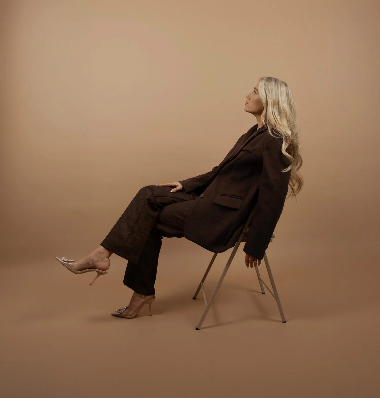 A woman with long blonde hair sitting sideways on a chair, wearing a dark suit and high heels with embellishments, against a plain beige background.