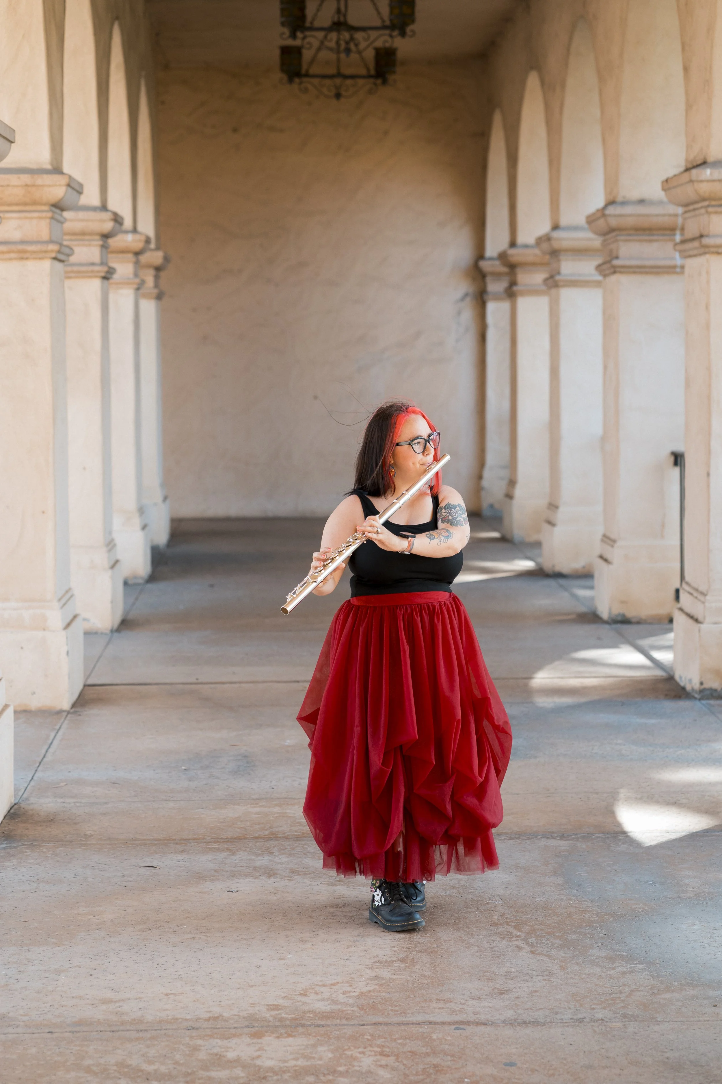 A woman with tattoos, glasses, and red hair playing a silver flute in an arched outdoor corridor with beige walls and columns, wearing a black top, red skirt, and black boots.