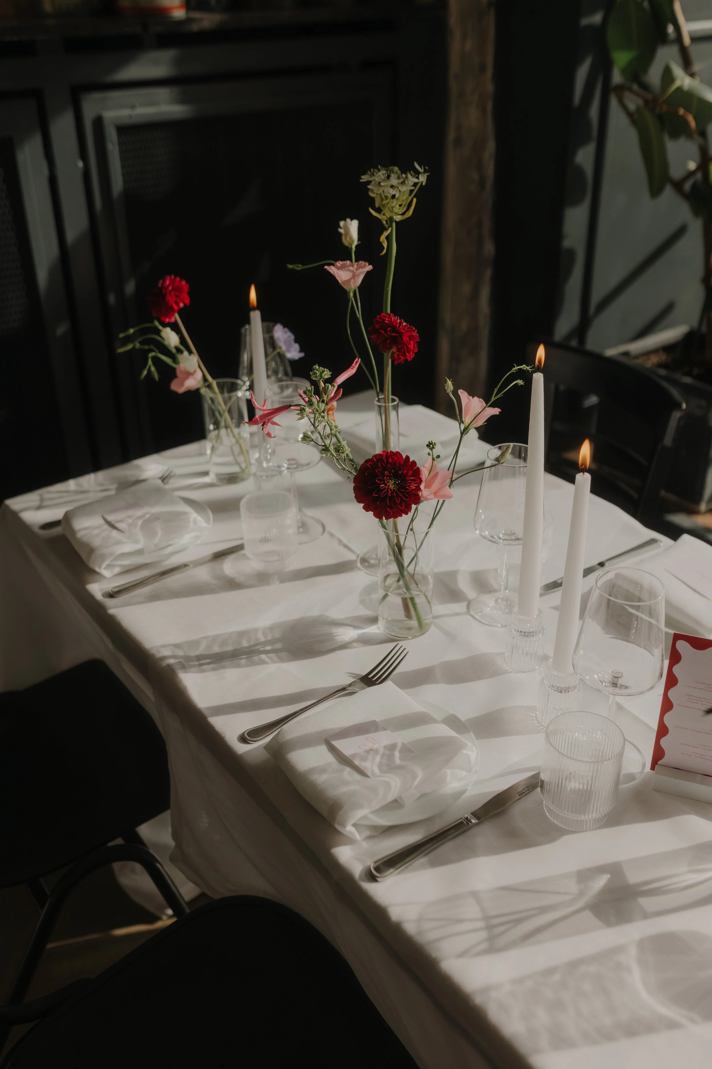 Elegant gedeckter Tisch mit weißen Tellern, Gläsern, Besteck und weißen Tüchern, geschmückt mit einer Blumenvasen mit roten und rosa Blumen sowie brennenden weißen Kerzen, in einem Raum mit dunklem Hintergrund.
