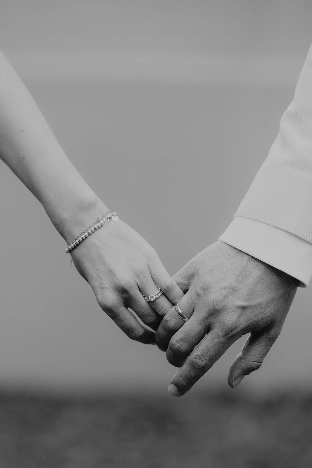 Ein schwarz-weiß Bild zeigt die Hände eines Paares, das sich hält. Die Frau trägt Ringe und Armbänder, der Mann trägt einen Anzug. Es wird eine emotionale Verbindung und Liebe dargestellt.