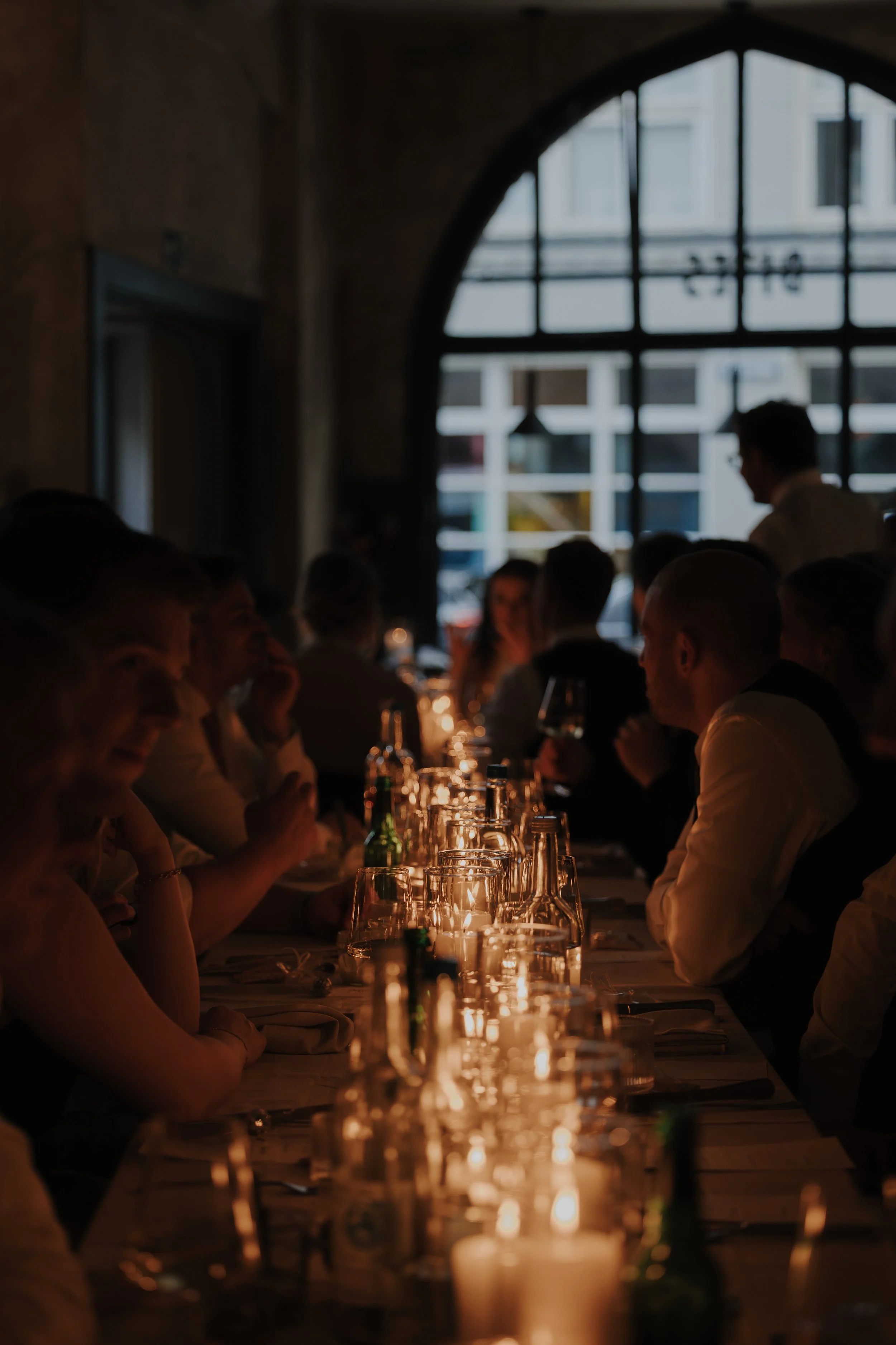 Eine dunkel beleuchtete Dinnerparty mit Menschen, die um einen langen Tisch sitzen, Kerzen und Getränke auf dem Tisch, im Hintergrund ein großes Fenster mit Sicht auf ein Gebäude.