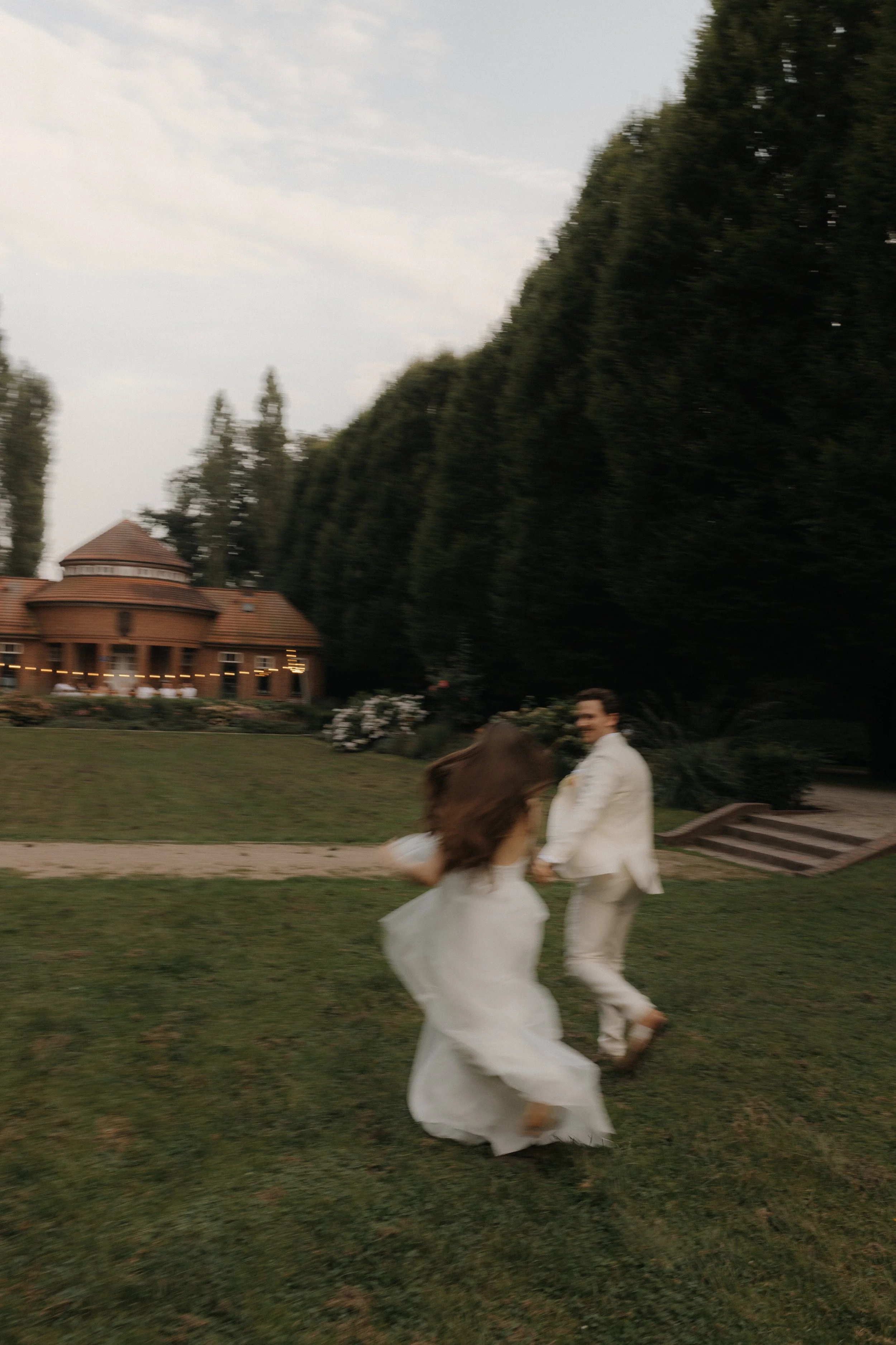Ein Mann und eine Frau tanzen im Freien auf einer grünen Wiese, im Hintergrund ein Haus mit roten Dächern und große Bäume.