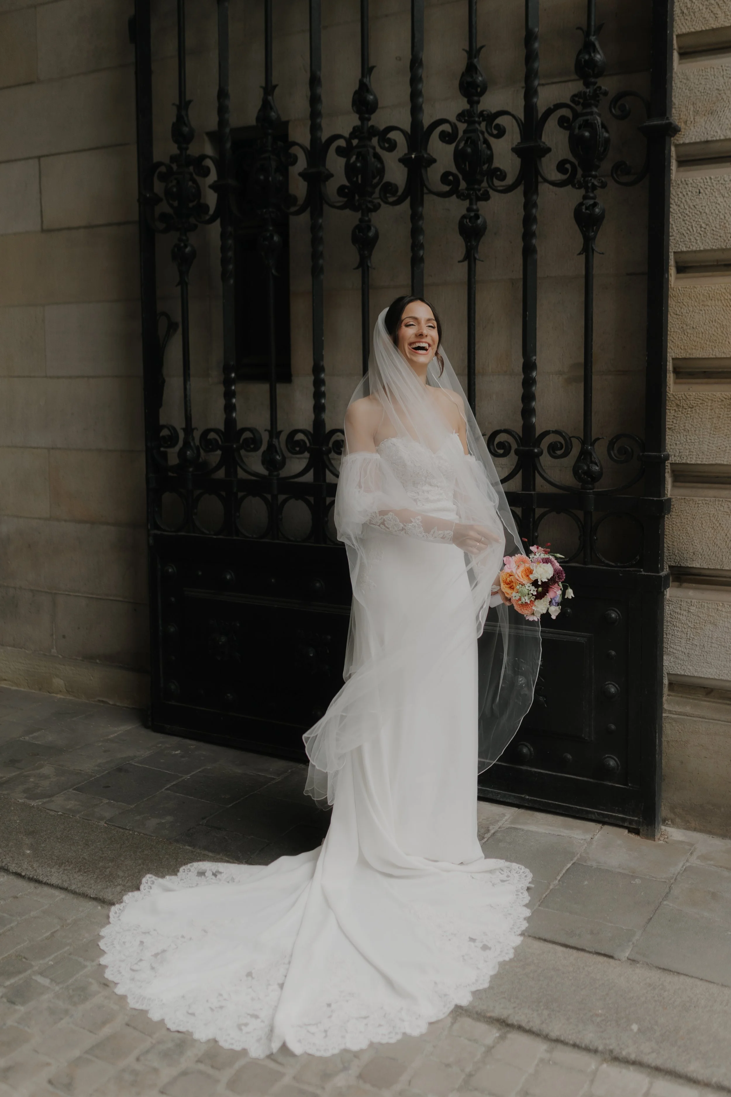 Braut mit weißem Kleid und Schleier, hält Bouquet, lacht vor schwarzem Gittertor auf Kopfsteinpflaster