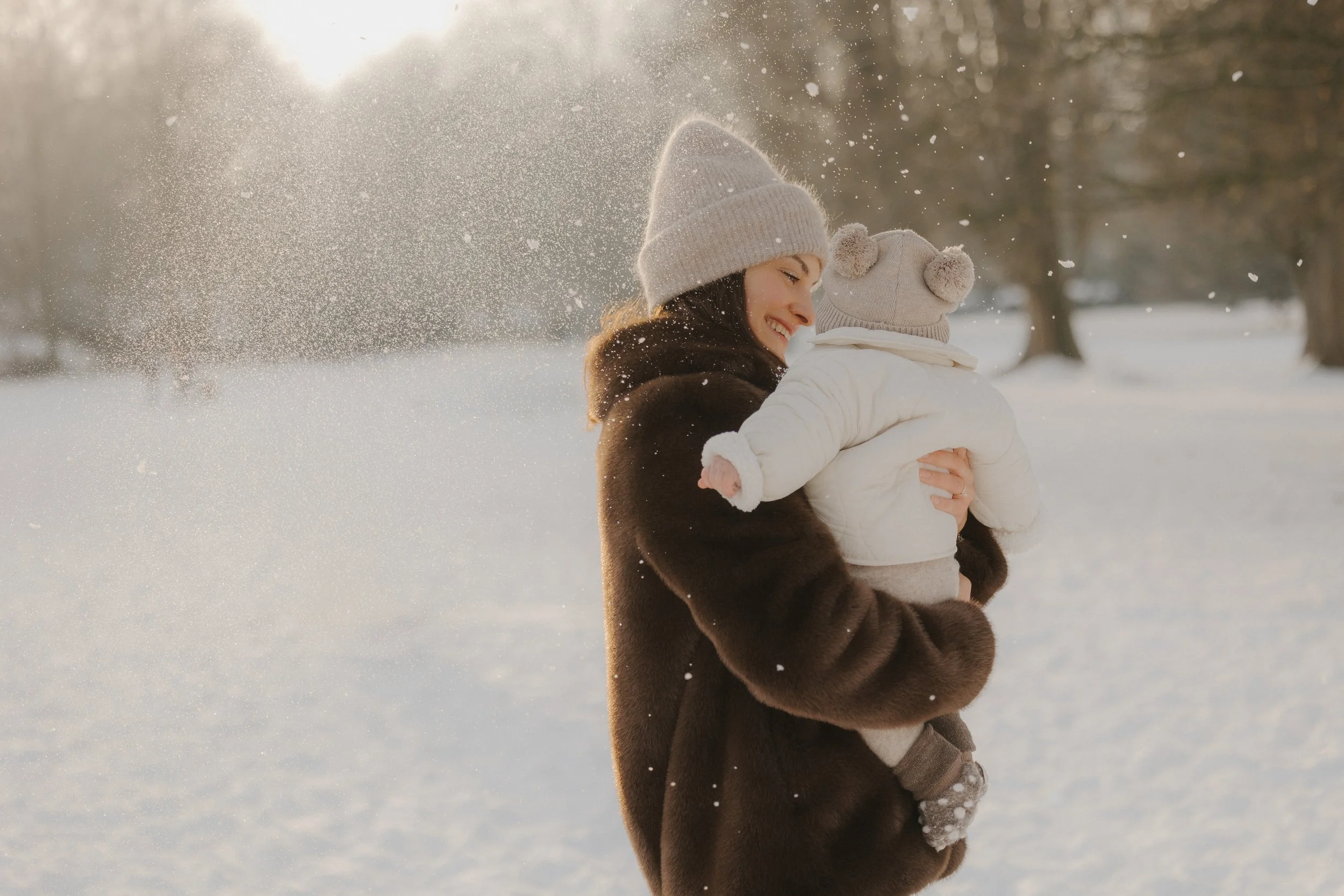 Eine Frau hält ein Kind in weiße Winterkleidung, beide lachen im schneebedeckten Park bei Sonnenlicht.