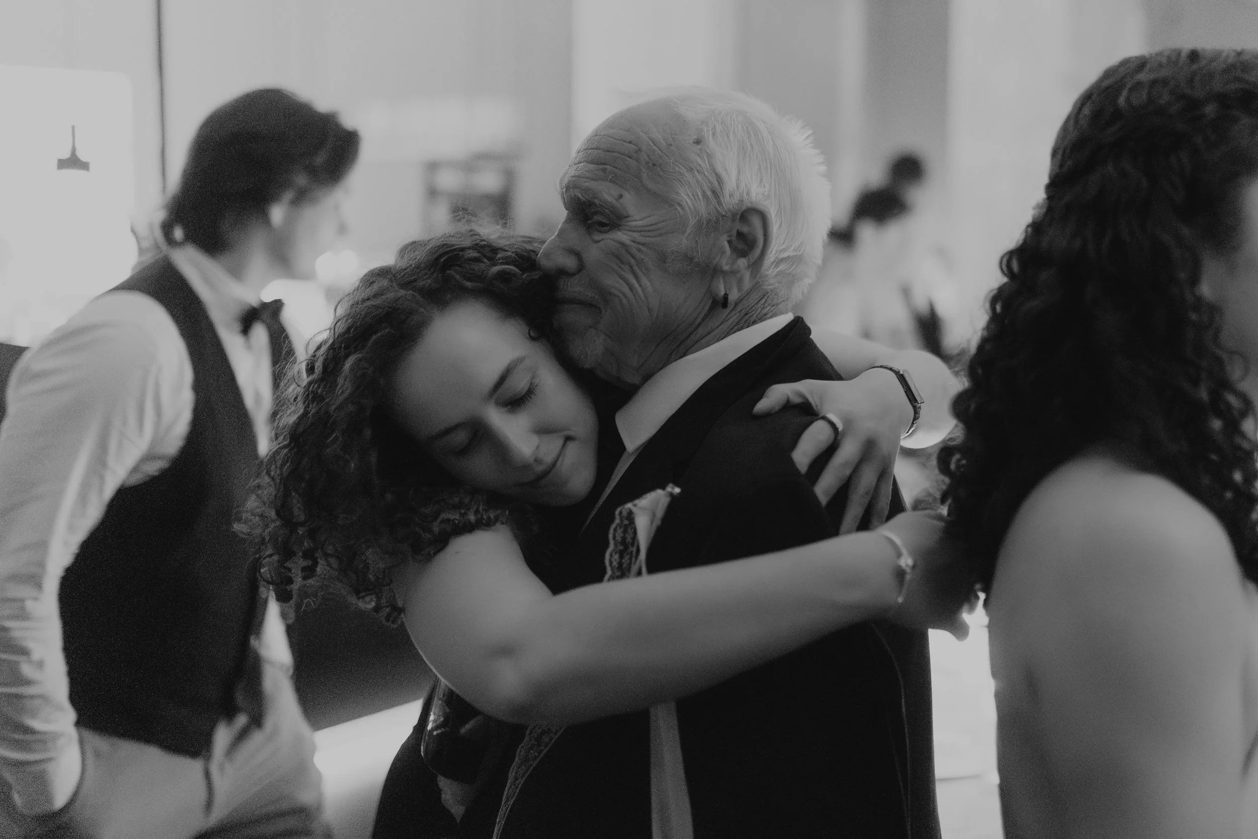 Zwei Frauen, die sich umarmen, im Hintergrund ein Mann im Anzug und eine Bar