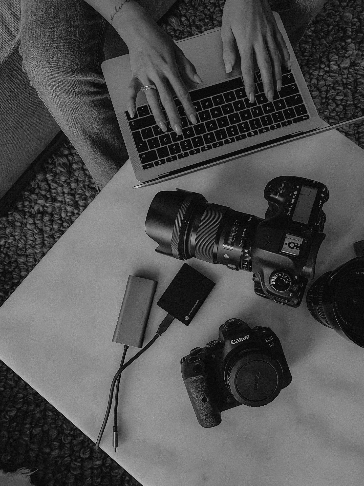 Eine Person tippt auf einer Laptop-Tastatur, auf einem weißen Tisch liegen eine professionelle Kamera, ein weiterer Kameraobjektiv, ein externer Akku, ein Kartenleser und ein zweiter Kamera. Der Hintergrund ist ein Teppich und ein Sofa.