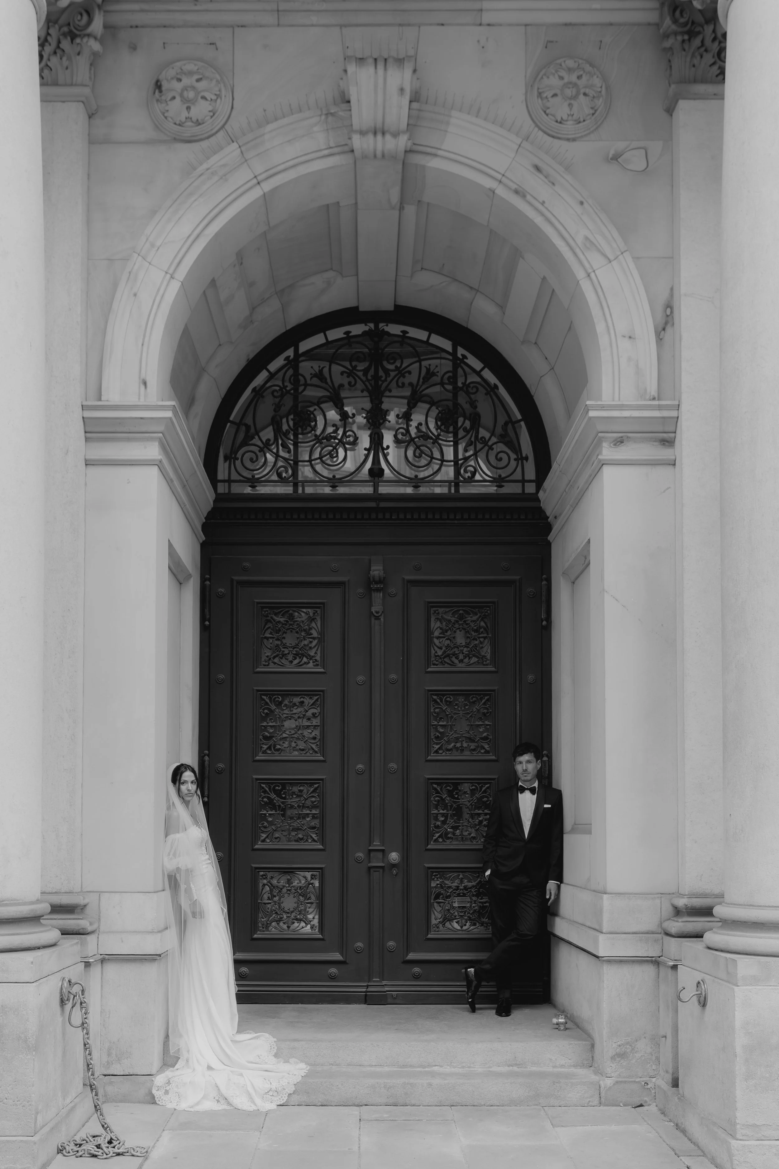 Ein schwarz-weiß Foto von einem Brautpaar, das vor einer großen, schwarzen, verzierten Tür an einer historischen Gebäude steht. Die Braut trägt ein langes, elegantes Kleid mit einem Schleier, der auf dem Boden liegt. Der Bräutigam trägt einen schwarz