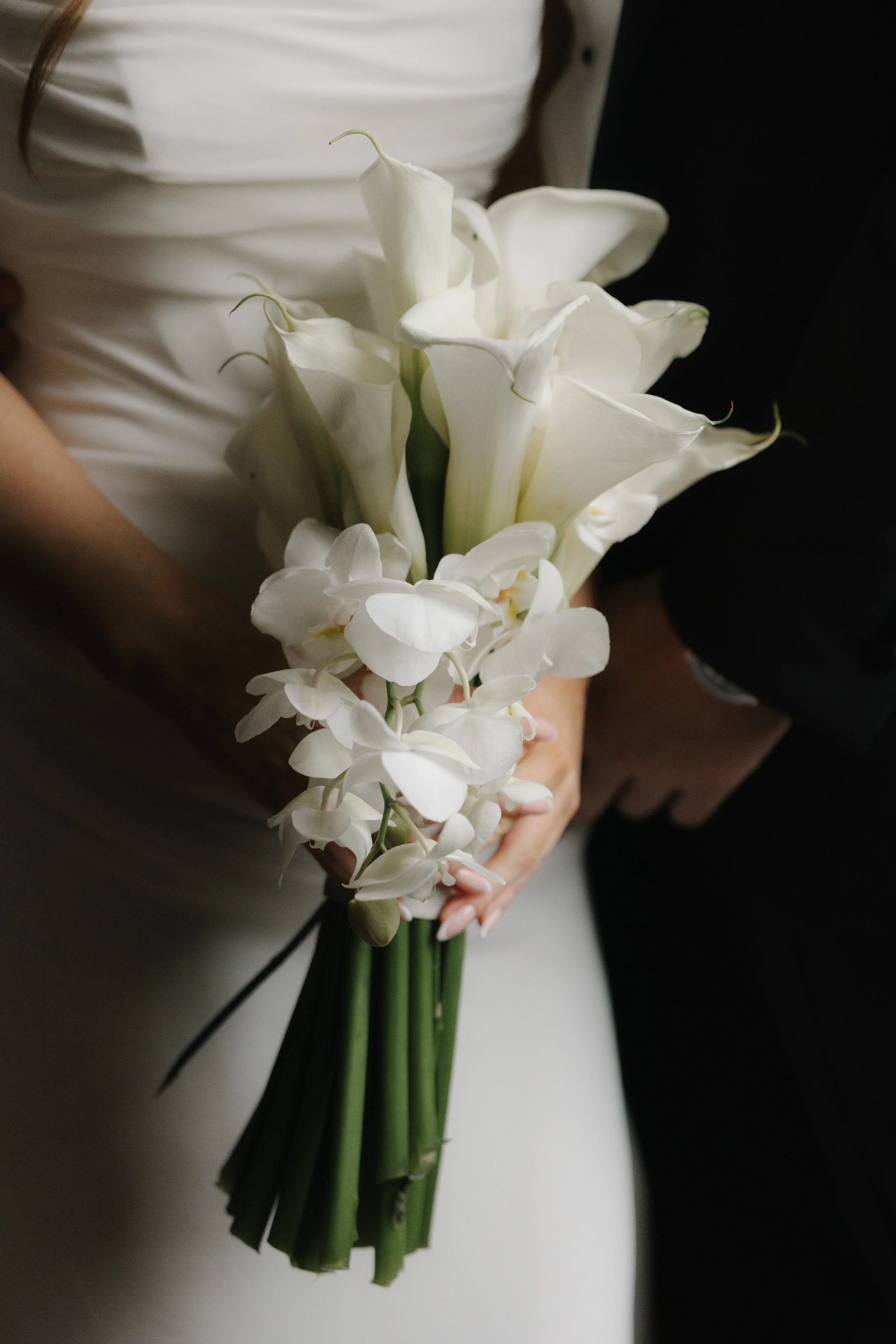 Blumenstrauß mit weißen Calla- und Orchideenblumen, gehalten von einer Person in formeller Kleidung.