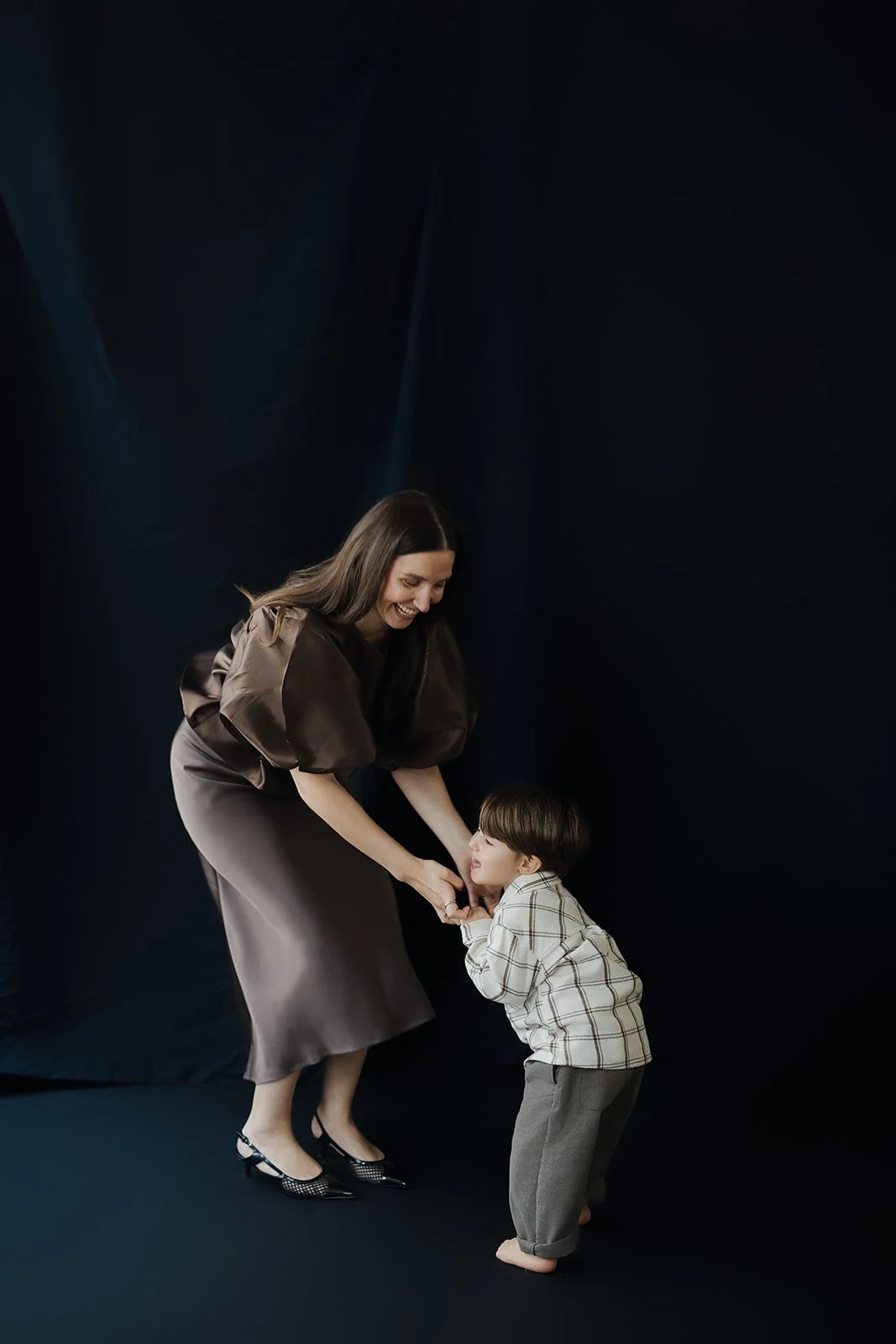 Frau und Kind, die sich lachend an den Händen halten, vor dunklem Hintergrund.